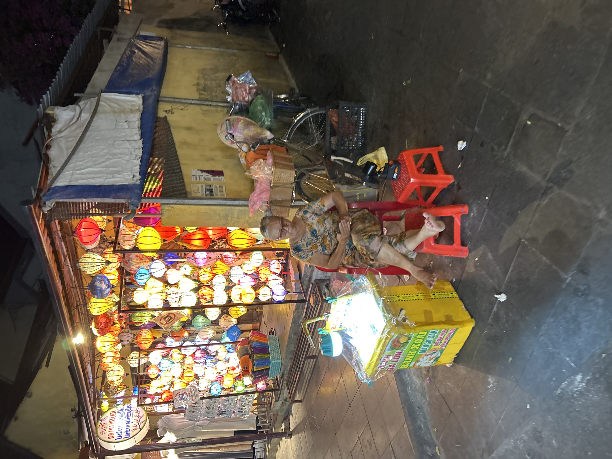 The image depicts a street scene at night in Hoi An Ward, Vietnam, characterized by a vibrant display of colorful lanterns. In the foreground, an elderly woman with glasses sits on a red plastic stool, her legs crossed. She is wearing a floral-patterned dress. To her left is a yellow, illuminated cart displaying food items and text in Vietnamese, including "BANH XOA" and "MANGO CAKE". A smaller red plastic stool is positioned to her right. Behind the cart, a bicycle is laden with baskets of goods and wrapped items. The background features a stall with numerous hanging lanterns of various colors and shapes, illuminated and creating a festive atmosphere. The stall also displays clothing on mannequins. The street is paved with dark tiles. The overall impression is that of a lively night market or street vendor scene.
