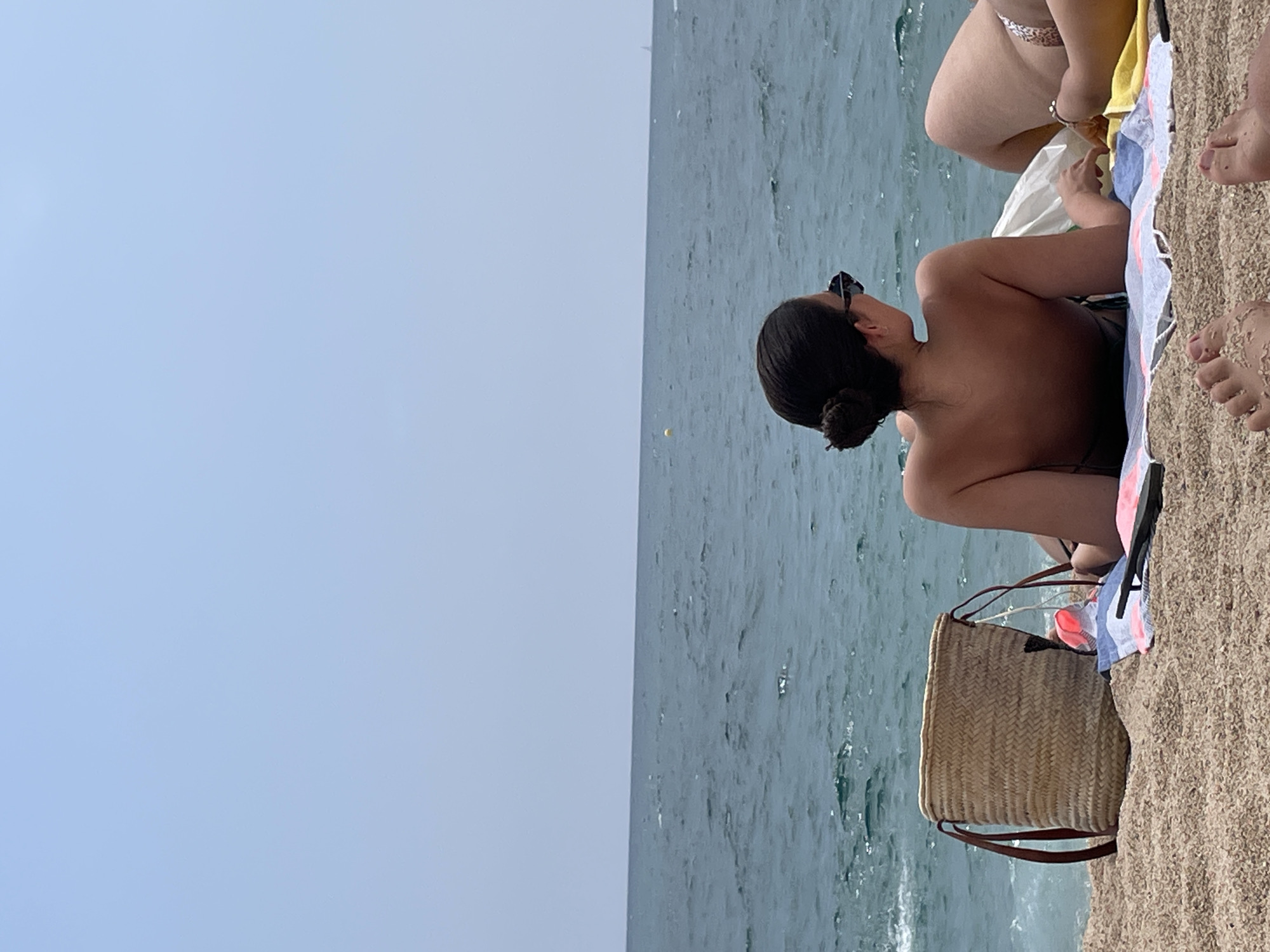 An overhead medium shot captures two individuals relaxing on a sandy beach in Canet de Mar, Spain, with the Mediterranean Sea in the background under a clear sky. A woman with her hair in a bun and wearing sunglasses is lying on a towel facing the ocean. To her right, another person's legs are visible, also resting on towels, one of which is yellow and the other is patterned. A woven straw bag with leather handles sits to the left of the woman, filled with a pink item. The sand is coarse and tan, and the water is a deep blue with gentle waves. The overall scene conveys a peaceful, sunny day at the beach.