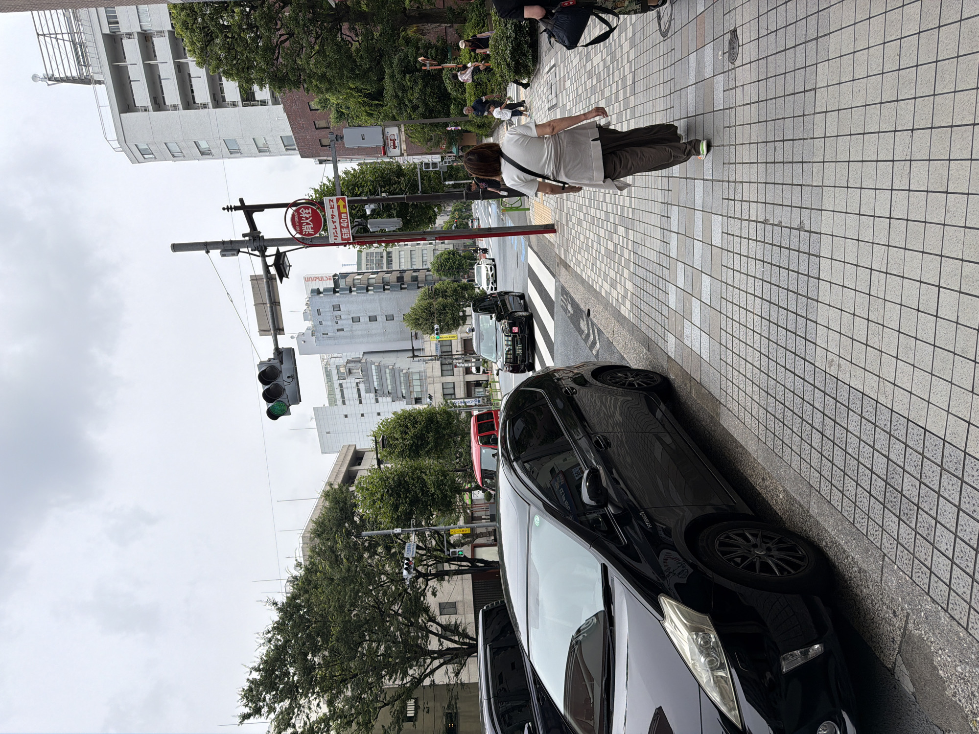 The image shows a street scene in Chuo, Japan, on an overcast day. In the foreground, a black hybrid car is parked on the left side of the frame, partially obscuring the sidewalk. The sidewalk is paved with light-colored square tiles and is wet, suggesting recent rain. A woman is walking away from the camera on the sidewalk, wearing a white t-shirt, brown pants, and brightly colored sneakers. Behind her, other pedestrians, including a child, are visible further down the sidewalk.
The street itself has multiple lanes of traffic. A black taxi and a red minivan are visible driving on the street, along with other vehicles in the distance. Traffic lights are visible, with one displaying a green signal. A fire hydrant sign is mounted on a pole, with Japanese text and an arrow indicating a direction. The background features several multi-story buildings of various architectural styles, interspersed with lush green trees. The overall impression is a typical urban environment in Japan on a cloudy day.
