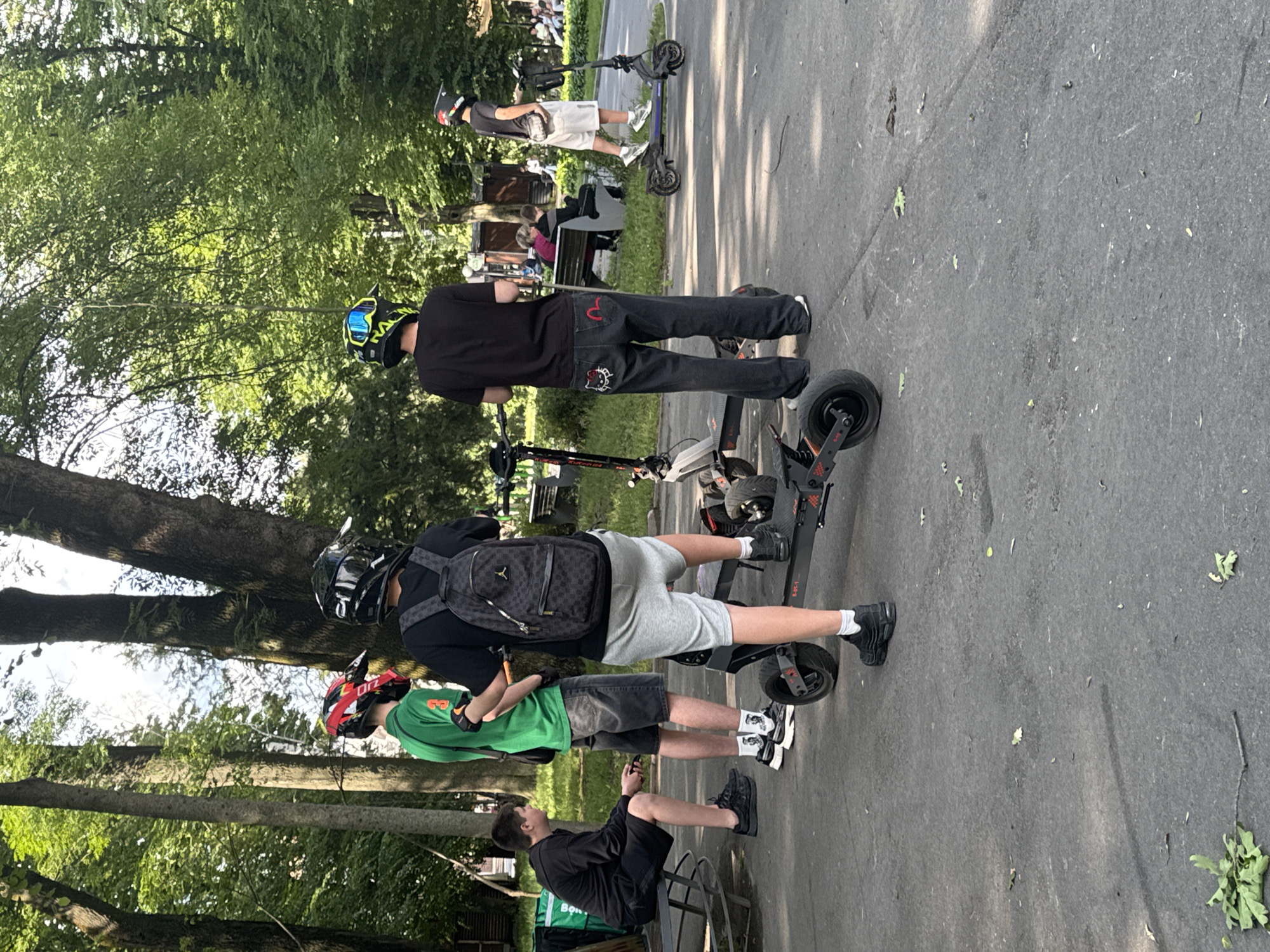 In Vinnytsia, Ukraine, a group of young people are gathered in a park, some wearing protective helmets and standing near electric scooters. The scene unfolds on a paved pathway, bordered by lush green trees and dappled sunlight. In the foreground, a young person in a black hoodie and shorts sits on a bench, holding a phone. Nearby, three other individuals stand, two of them wearing helmets and the third in a black t-shirt and dark jeans. They are positioned next to several electric scooters, suggesting an outing or a pause in their activities. Further back, other park-goers can be seen relaxing on benches amidst the trees. The atmosphere appears casual and recreational, typical of a sunny day in a city park.