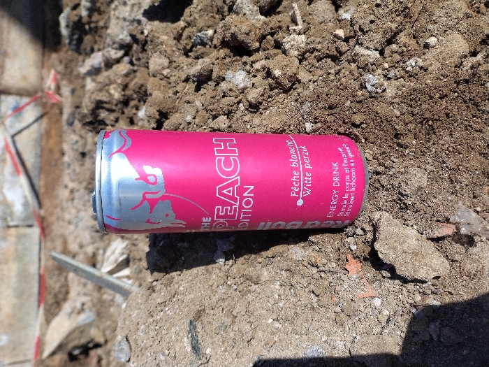 A pink and silver Red Bull "The Peach Edition" energy drink can, featuring text in French ("Pêche blanche") and Dutch ("Witte perzik"), stands upright on a pile of loose, dry brown soil mixed with small stones and debris. The can's surface shows a prominent silver bull logo. Bright sunlight illuminates the scene, casting a shadow to the lower left of the can. In the blurred background, piles of similar earth and fragmented construction materials are visible, alongside what appears to be a concrete structure and a red and white striped barrier tape. The scene is located in Crauthem, Luxembourg.