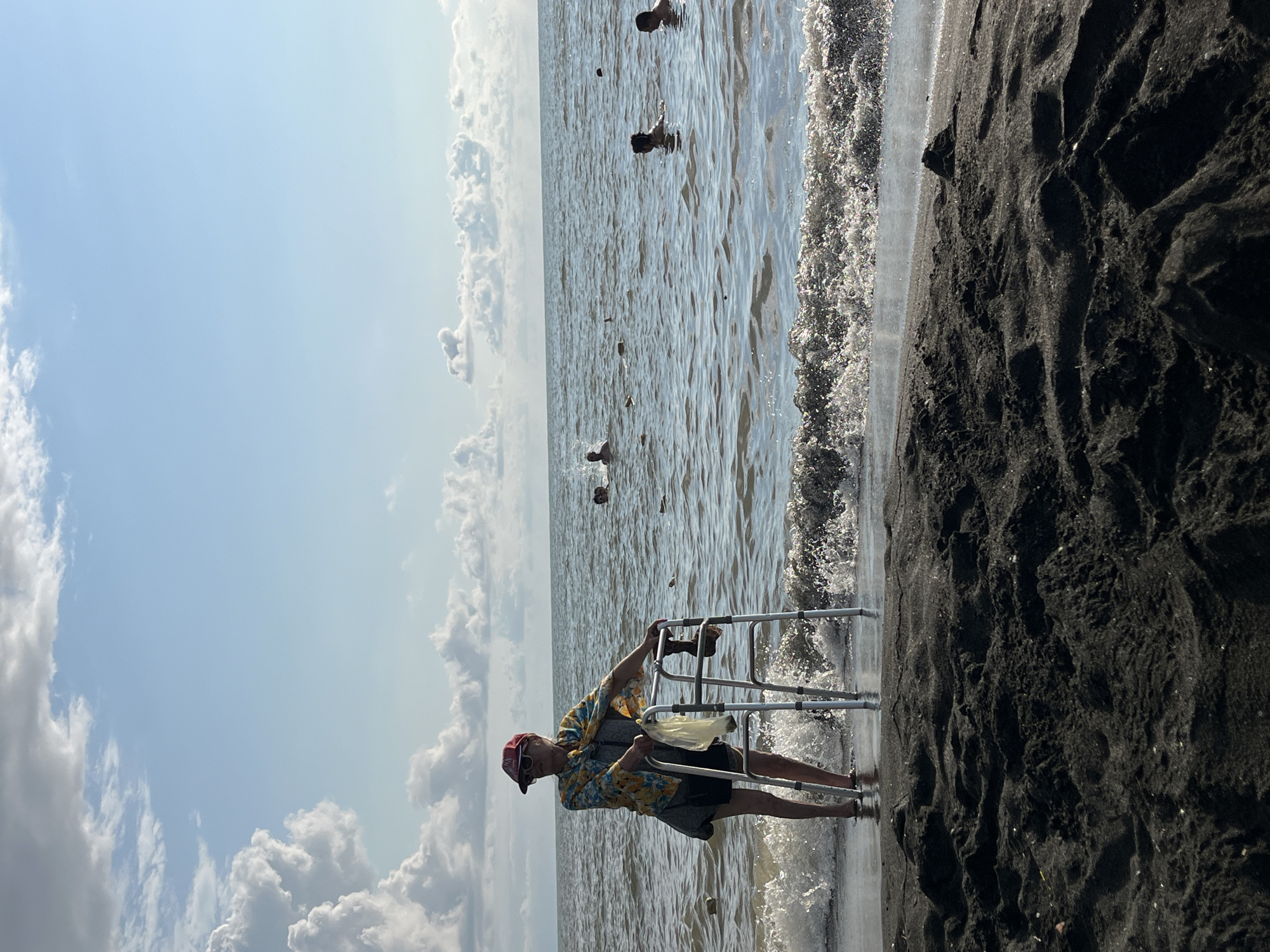 A person stands on a sandy beach with a walker in the shallow water of the sea. The person is wearing a colorful short-sleeved shirt, dark shorts, sunglasses, and a red baseball cap. They are holding the walker with both hands, and a plastic bag is hanging from the walker. The sea is calm with some small waves breaking on the shore, and there are several people swimming in the background. The sky is blue with scattered clouds. The sand on the beach is dark and appears wet near the water. The scene is located on the Black Sea coast in Ureki, Georgia, known for its magnetic sand beaches.