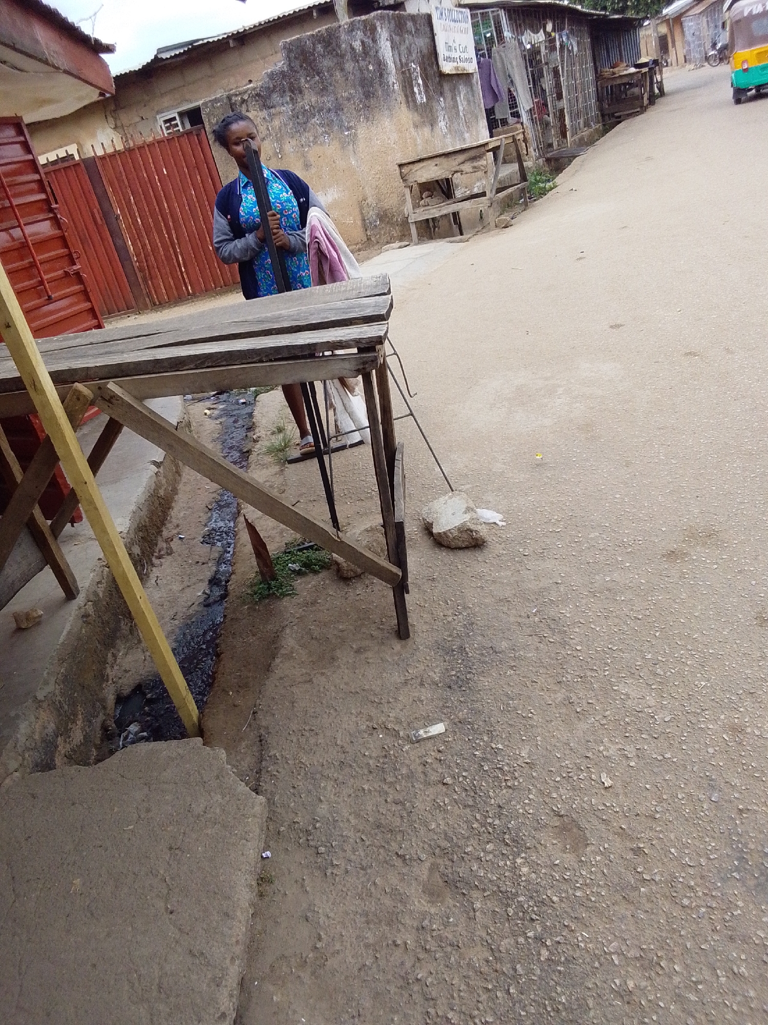 The image depicts a vibrant street scene in Jos, Nigeria, characterized by an unpaved, gravelly road and simple, functional architecture. In the foreground, a makeshift wooden stall or table structure stands on the left, partially obscuring a narrow, dark gutter filled with murky water and debris that runs alongside the street. The road extends into the background, where more basic buildings and stalls line its edges, with a yellow and green auto-rickshaw visible in the distance.
A woman, appearing to be a local resident or vendor, stands next to the wooden structure on the left. She is dressed in a blue patterned top over a grey long-sleeved garment, and a light-colored cloth is draped over her arm. Her dark hair is pulled back, and she holds a dark, rectangular object, possibly a metal bar, near her face. She wears sandals, and her expression appears calm. Behind her is a red corrugated metal gate.
Notable details include a white sign on a building further down the street that clearly reads "TIM'S COLLECTION," "DRESSES OF A LADY," "TIM'S CUT," and "BARBING SALOON," indicating local businesses. A large white rock and some litter are visible on the gravelly ground. The diffused lighting suggests it is daytime, possibly under an overcast sky, as there are no harsh shadows. The overall setting portrays a typical, bustling street environment in a semi-urban or rural area of Nigeria.