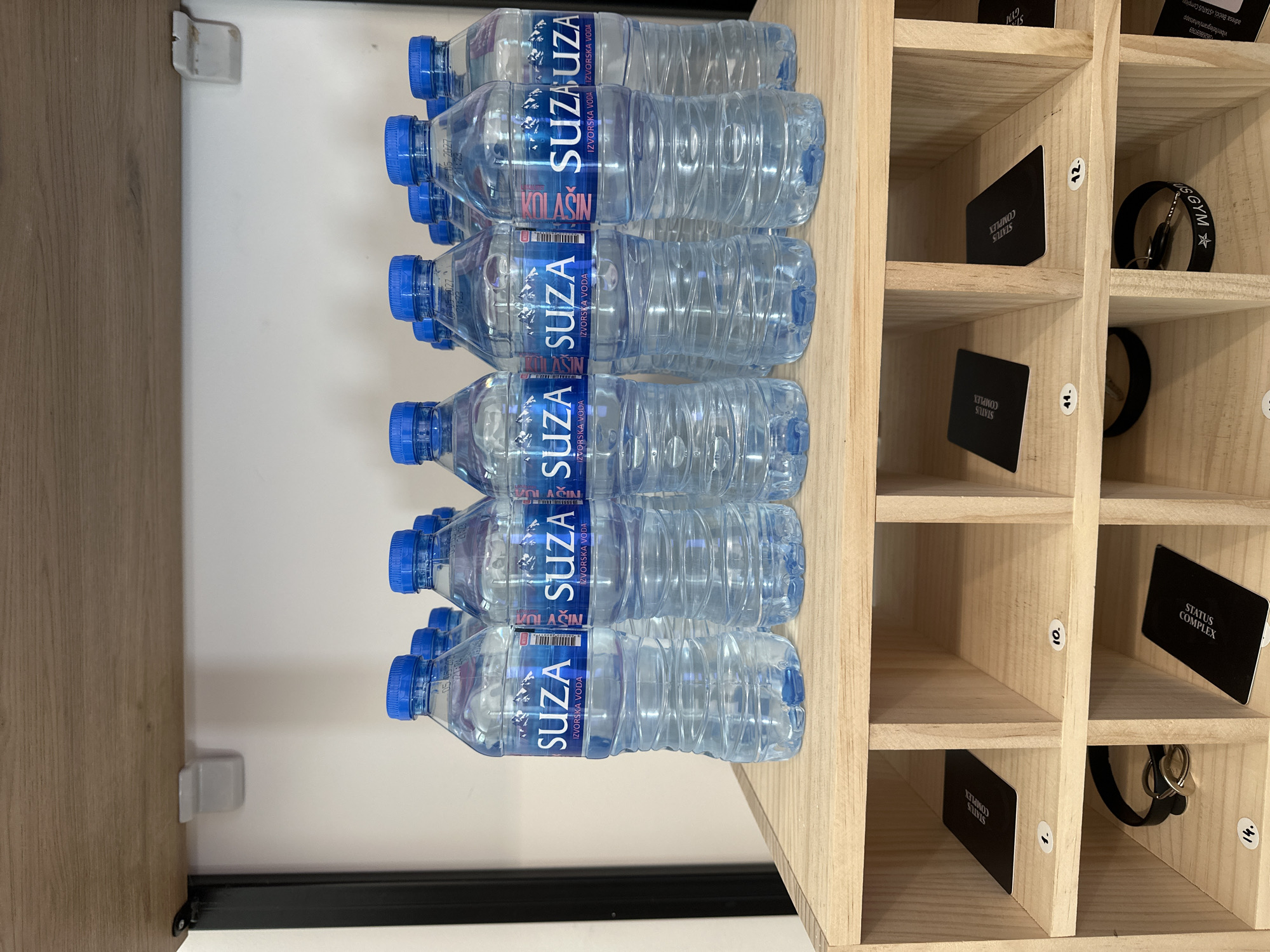 In Boreti, Montenegro, a wooden shelving unit displays several bottles of Suza Izvorska Voda water. The bottles have blue caps and clear plastic with blue and white labels featuring mountains and the brand name. Below the bottles, the shelving unit has individual compartments. Some compartments contain black cards with white text reading "STATUS COMPLEX". Other compartments hold keychains with black bands and metal rings. The shelves are numbered with white circular stickers: "1.", "10.", "11.", "12.", and "14.". The environment appears to be indoors, with a neutral-colored wall in the background and a wooden shelf above.