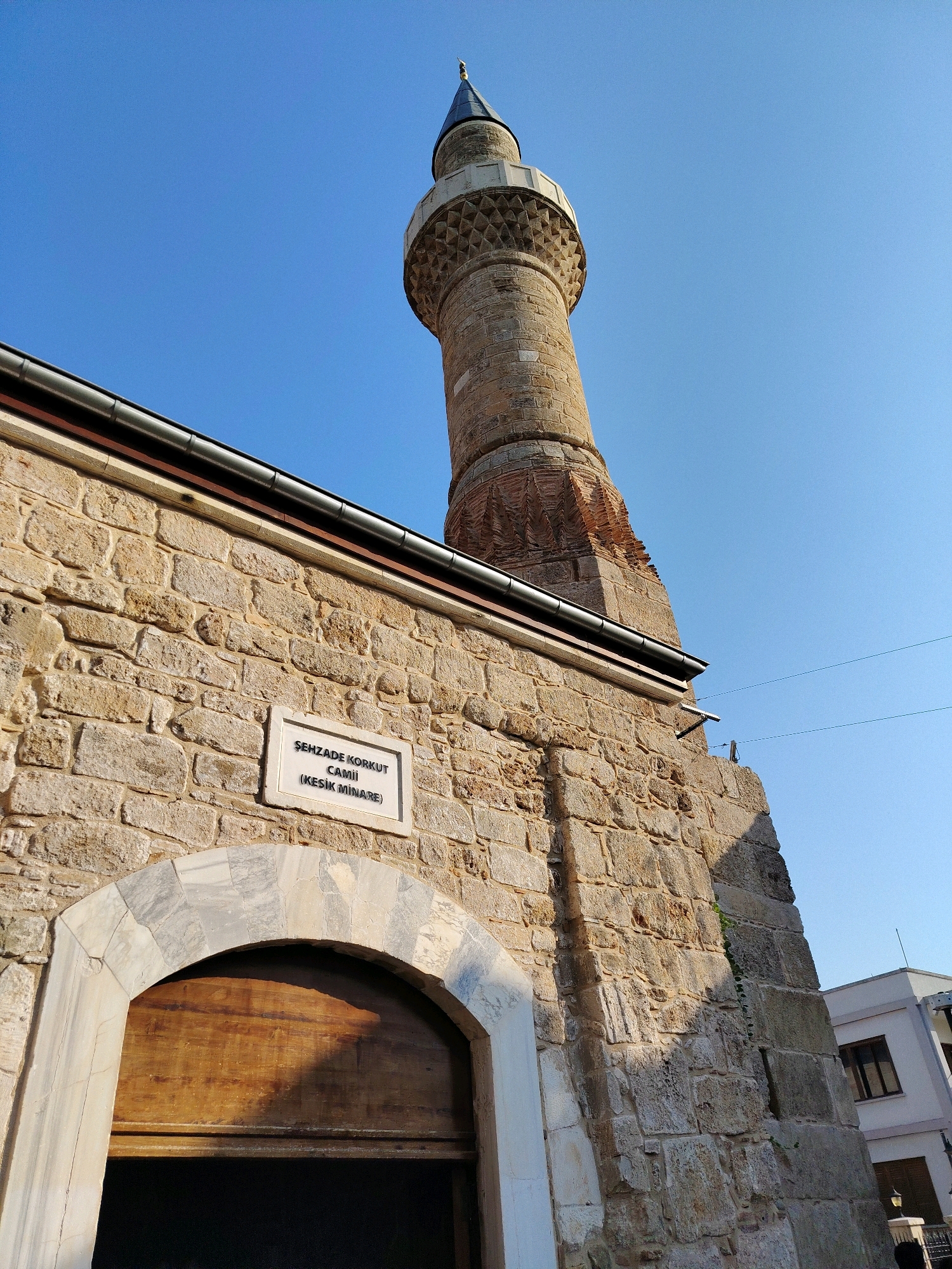 A stone-built mosque minaret and entrance with a wooden door are visible under a clear blue sky. A sign on the stone wall reads "ŞEHZADE KORKUT CAMII (KESİK MİNARE)". The minaret is tall and has a stone shaft with intricate geometric patterns near the top. The mosque entrance features a marble archway and a large wooden door. Adjacent to the mosque, a modern building with white walls and brown window frames is partially visible. The scene is likely in Antalya, Turkey, as suggested by the "Kesik Minare" or "Broken Minaret" reference, which is a prominent historical landmark in the city.