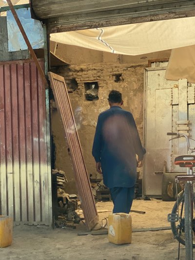 A man in dark blue traditional clothing walks into a building in Kandahar, Afghanistan. The structure appears to be a workshop of some kind with metal doors, concrete floors, and a brick wall with a rectangular opening. A faded white tarp serves as a ceiling or awning. A bicycle is visible to the right, and yellow plastic containers are on the floor. A wooden panel leans against a red metal door. The overall condition of the scene indicates a working-class environment.