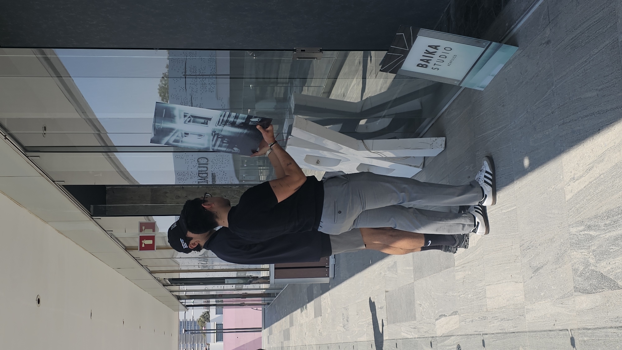 Two adult males are positioned outdoors on a light grey tiled walkway next to a modern building with extensive glass facades, in Puebla, Mexico. The man on the right, wearing glasses, a black t-shirt, light grey trousers, and white Adidas Samba sneakers, holds a large rectangular grayscale architectural print depicting a modern interior space. He is presenting or affixing the print against the reflective glass window. Tattoos are visible on his forearms. To his left, a second male, clad in a black baseball cap, black long-sleeved shirt, grey shorts, and dark athletic shoes, stands facing the glass, observing the action. A large, abstract, white sculptural object rests on the ground between the men and the window. Adjacent to this object, a freestanding vertical sign displays "BAIKA STUDIO ACAPULCO" with a blue-green gradient. The glass facade reflects a clear sky, distant urban buildings, and inverted text, including "CIUDAD." Above the glass, a red square sign with a white fire extinguisher icon is visible. The scene is illuminated by bright sunlight, casting distinct shadows on the pavement.