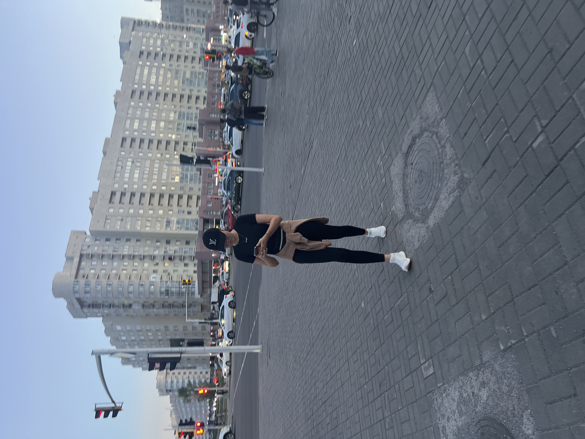 The image depicts a street scene in Astana, Kazakhstan, with a woman in the foreground walking while looking at her phone. She is wearing a black t-shirt, black leggings, white sneakers, and a tan sweater tied around her waist. She is also wearing a black baseball cap with a white logo. Behind her, a busy intersection is visible with cars and traffic lights. Several apartment buildings line the street in the background. The sky is a clear, light blue, indicating it is either early morning or late afternoon. The ground is paved with grey interlocking bricks. In the mid-ground, other people are seen walking and riding bicycles, and several cars are stopped at the intersection. The overall impression is of a typical urban environment in Kazakhstan's capital.