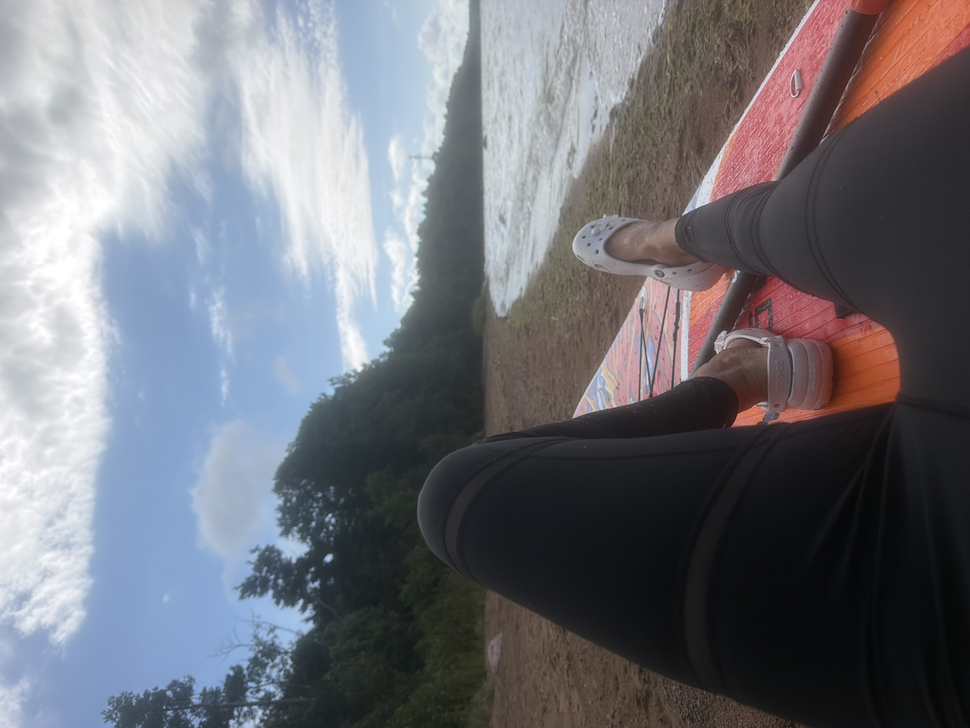 The image shows a person's legs, wearing black leggings and white Crocs, resting on an orange paddleboard on a sandy beach. The paddleboard has some colorful graphics and a black paddle is visible. The beach is bordered by a dense, dark green forest that slopes upwards. The sea is to the right, with choppy, light-colored waves and some seaweed visible along the shoreline. The sky is blue with scattered white clouds, some of which are wispy and thin, suggesting a partly cloudy day. There are no other people or significant structures visible in the immediate vicinity. The overall scene depicts a relaxed moment by the water, likely in a natural, undeveloped area. The image does not provide any specific details to identify the location as Vyazy, Russia.