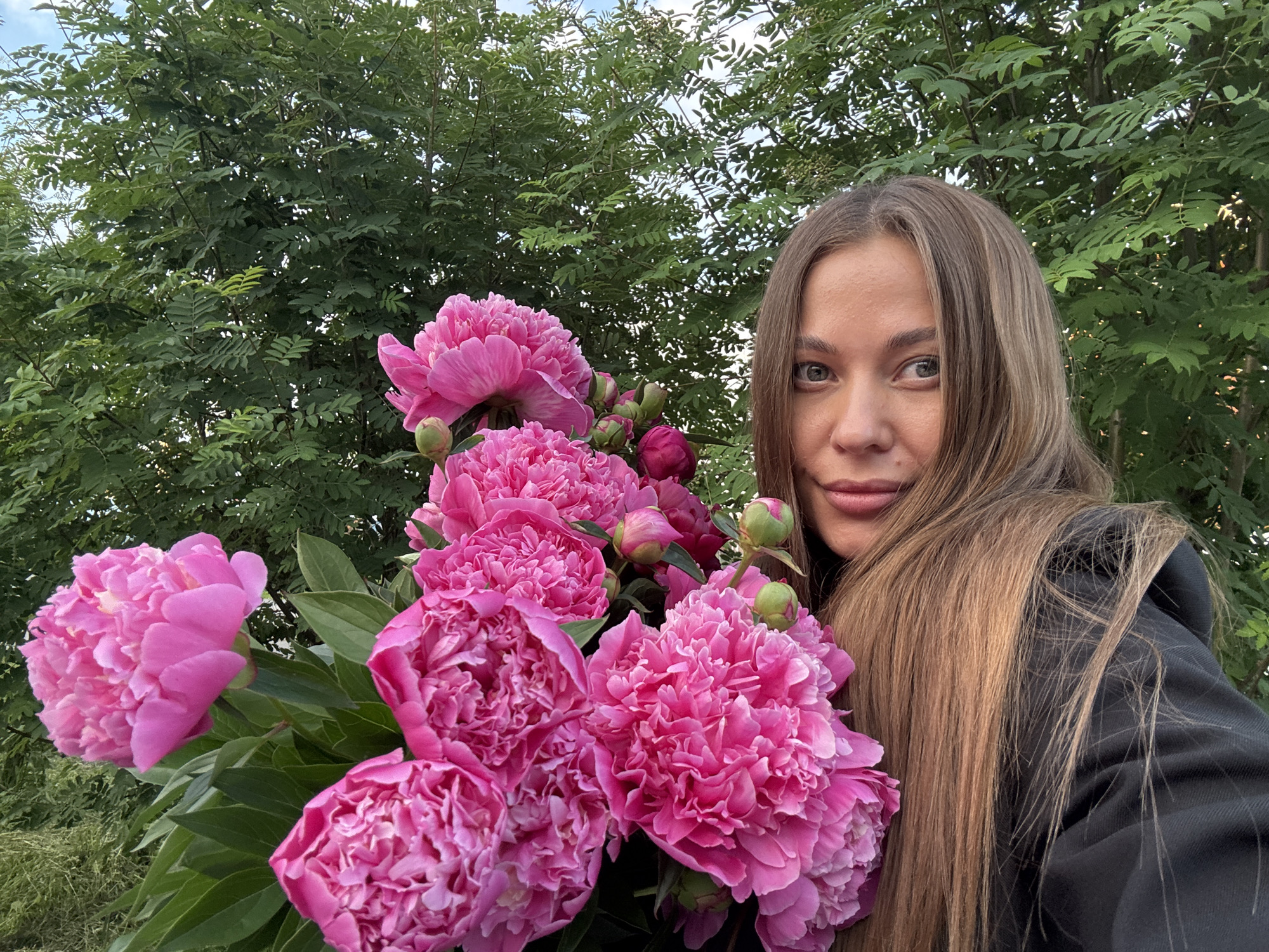 A woman with long, light brown hair is holding a bouquet of bright pink peonies in front of a backdrop of green trees and foliage. She is wearing a black hoodie and is looking towards the camera with a slight smile. The image appears to be taken outdoors.