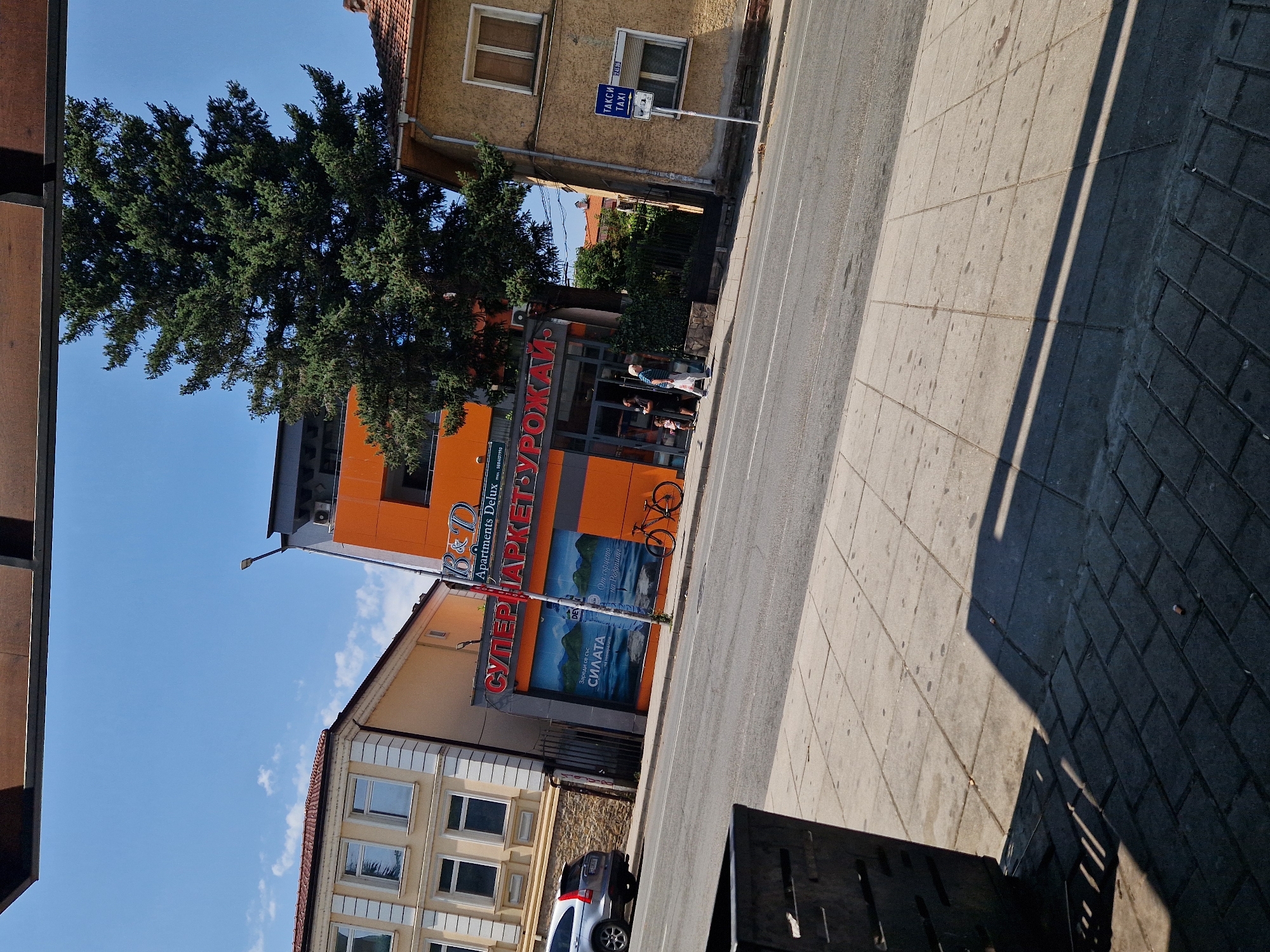The image shows a street scene in Lovech, Bulgaria, on a sunny day. In the foreground, a grey concrete sidewalk is visible, with a dark metal trash receptacle on the left. The sidewalk transitions to a road, where a silver SUV is parked on the left. Across the street, there are several buildings. The most prominent is a modern, orange and grey building with a sign reading "B&D Apartments Delux" and "СУПЕРМАРКЕТУРОЖАЙ". A large evergreen tree stands behind this building. To the right of the orange building is a yellow building with a red-tiled roof, displaying a blue sign with white text that reads "ТАКСИ TAXI". Several people are seen walking on the sidewalk in front of the supermarket, and a bicycle is parked nearby. The sky is clear and blue.