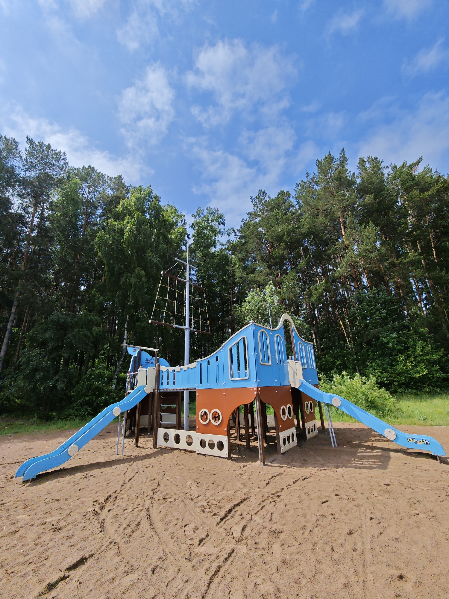 A vibrant pirate ship-themed playground takes center stage in this outdoor scene, set on sandy ground with a dense forest background under a bright blue sky. The playground structure is predominantly blue and brown, resembling a ship with a tall mast featuring a rope climbing net. Two long, curved blue slides extend from the ship's sides down to the sandy surface, one on the left and one on the right.
The sandy foreground shows various footprints and disturbances, indicating it's a well-used area. Numerous round "00" or "OO" shaped cutouts are visible along the lower part of the ship's structure, possibly serving as decorative elements or small windows. On the blue slide to the right, a small white logo or text that reads "AJAX P" can be discerned.
Behind the playground, a thick expanse of tall evergreen and deciduous trees creates a lush, natural backdrop. The sky above is a clear, bright blue with scattered, wispy white clouds, suggesting a sunny and pleasant day. Sunlight casts distinct shadows from the playground equipment onto the sand. No people are visible in the image. The setting appears to be a recreational park or a natural area, consistent with the provided location context of Chaikovsky, Russia.
