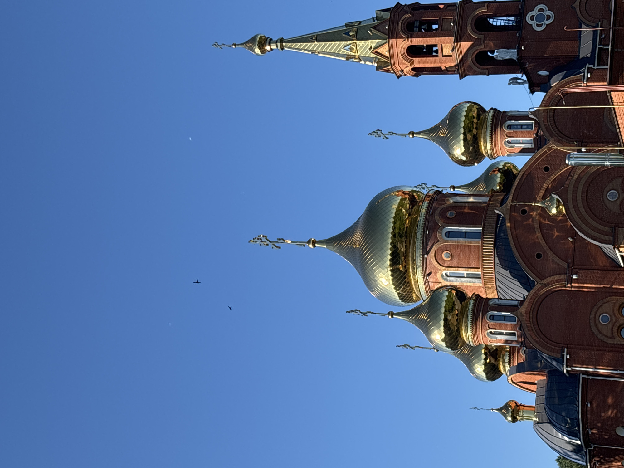 The image shows a red brick Orthodox church with golden domes and a tall bell tower, against a clear blue sky. Several birds are visible in flight against the sky. The architecture suggests a building of religious significance, likely the Cathedral of the Transfiguration in Chaikovsky, Russia. The sun is casting shadows on the building, indicating it is likely daytime. There are no people visible in the image.
