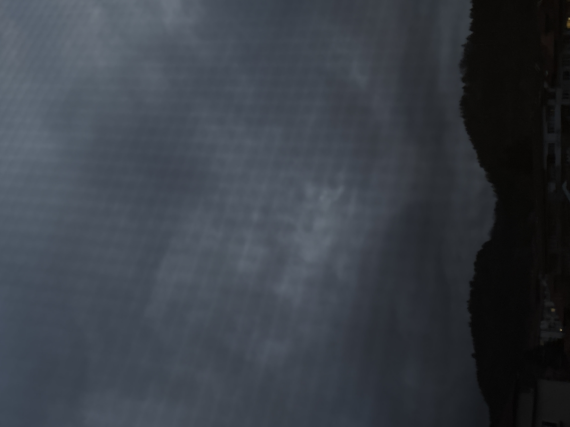The image captures a panoramic view during nighttime or late evening in the city of Troyan, Bulgaria. The upper two-thirds of the frame are filled with a dark, heavily overcast sky, exhibiting subtle variations in cloud density. A faint grid-like artifact pattern is observable across the sky. The lower third of the image features the dark, silhouetted outlines of forested hills or mountains. Below these hills, in the foreground, faint forms of urban buildings are visible. Multiple windows within these structures emit warm, artificial light, indicating interior illumination. No individuals, explicit actions, or specific subjects beyond the landscape and distant buildings are discernible.