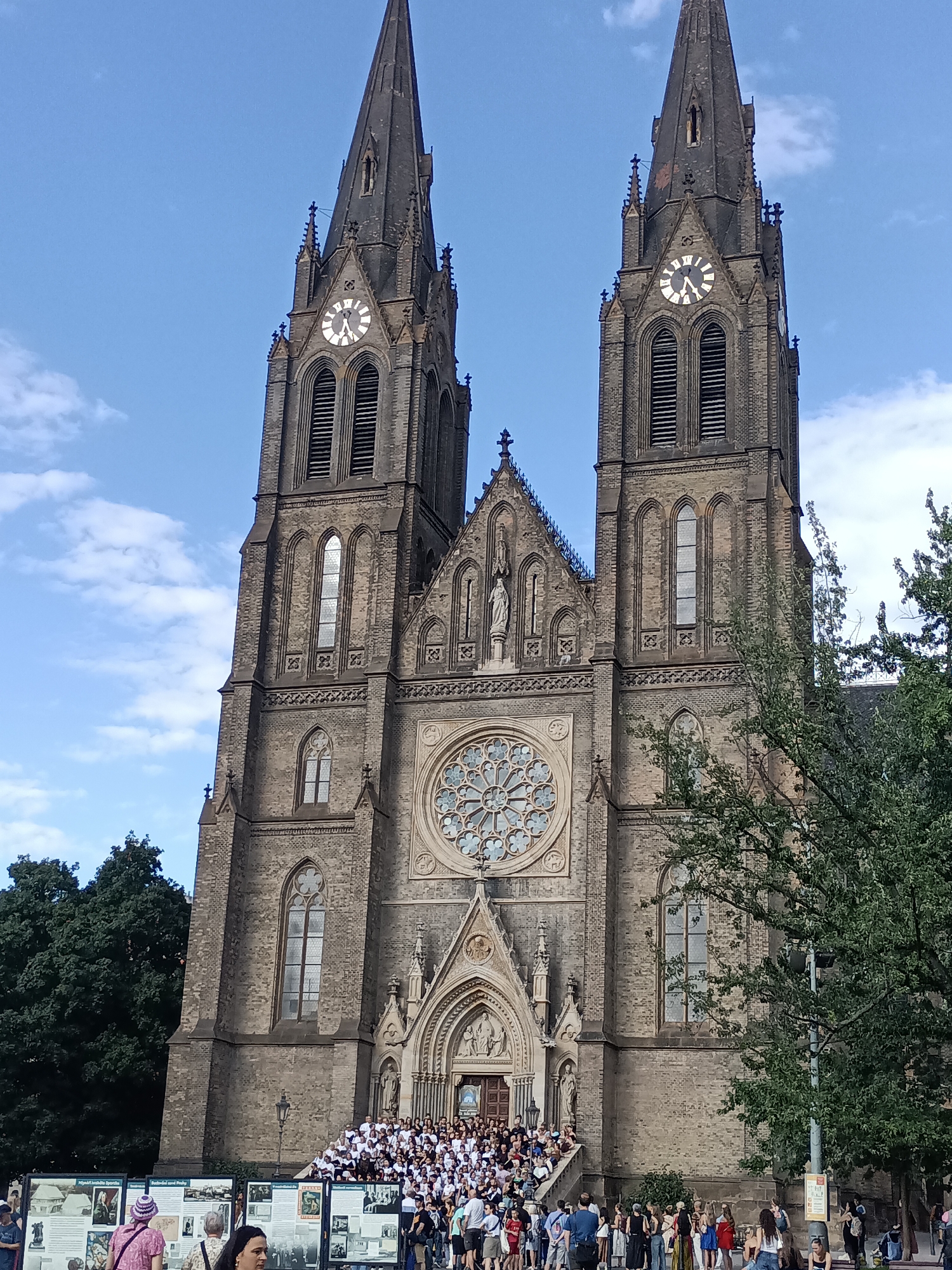 This is a medium-angle, full-body shot of the Basilica of Saint Peter and Saint Paul in Prague, Czechia, captured on a bright, partially cloudy day. The imposing Neo-Gothic facade of the church dominates the frame, with its twin spires reaching towards the sky. A clock is visible on the right spire, showing the time as approximately 12:50. In front of the church entrance, a large crowd of people, mostly dressed in white, are gathered, with some seated on the steps leading up to the basilica. Several individuals are standing and looking at the church or interacting with each other. In the foreground, to the left, are informational display boards with images and text, and more people are visible browsing them. To the right, lush green trees partially obscure the right side of the church, and a lamppost stands on the sidewalk. The overall atmosphere appears to be a public gathering or event in front of a prominent historical landmark.