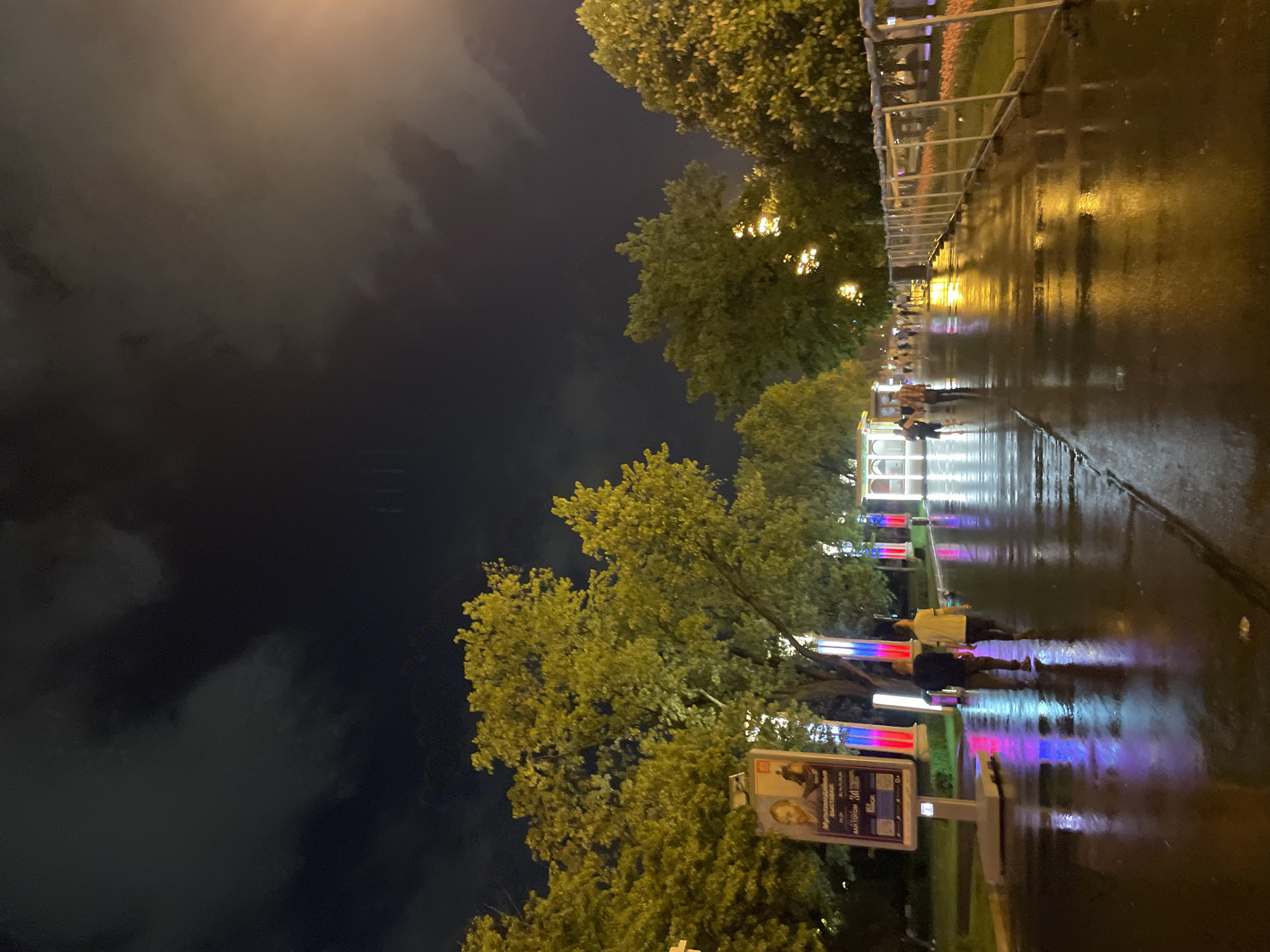 The image depicts a nighttime scene in Moscow, Russia, specifically on a rain-slicked pedestrian walkway illuminated by colorful, vertical light installations. Trees line the walkway. Several pedestrians are visible, walking along the path. On the left side, a poster is displayed, suggesting a local event or exhibition. The sky above is dark, with the implication of overcast conditions.