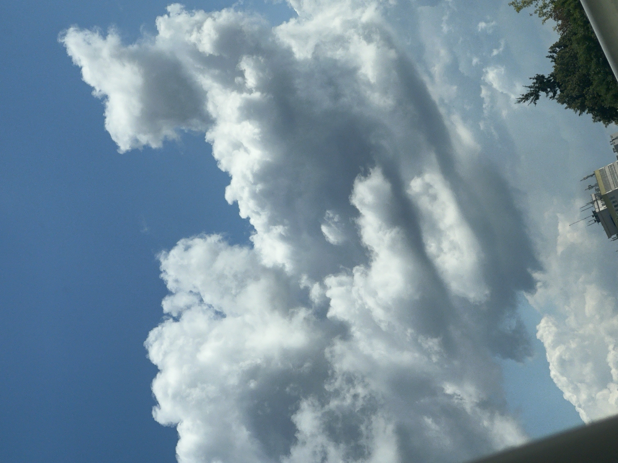 The image shows a sky with dramatic clouds, likely captured from a moving vehicle. The clouds are predominantly white and grey, with some areas appearing darker, suggesting varying thickness and light penetration. The sky is blue, indicating daylight. In the lower part of the image, a building with antennas and a tree line are visible. The building appears to be modern with a light-colored facade and multiple levels. The tree line consists of dense green foliage. The overall impression is that of an urban or suburban landscape under a cloudy sky. The specific location within Warsaw, Poland cannot be determined from the provided image.