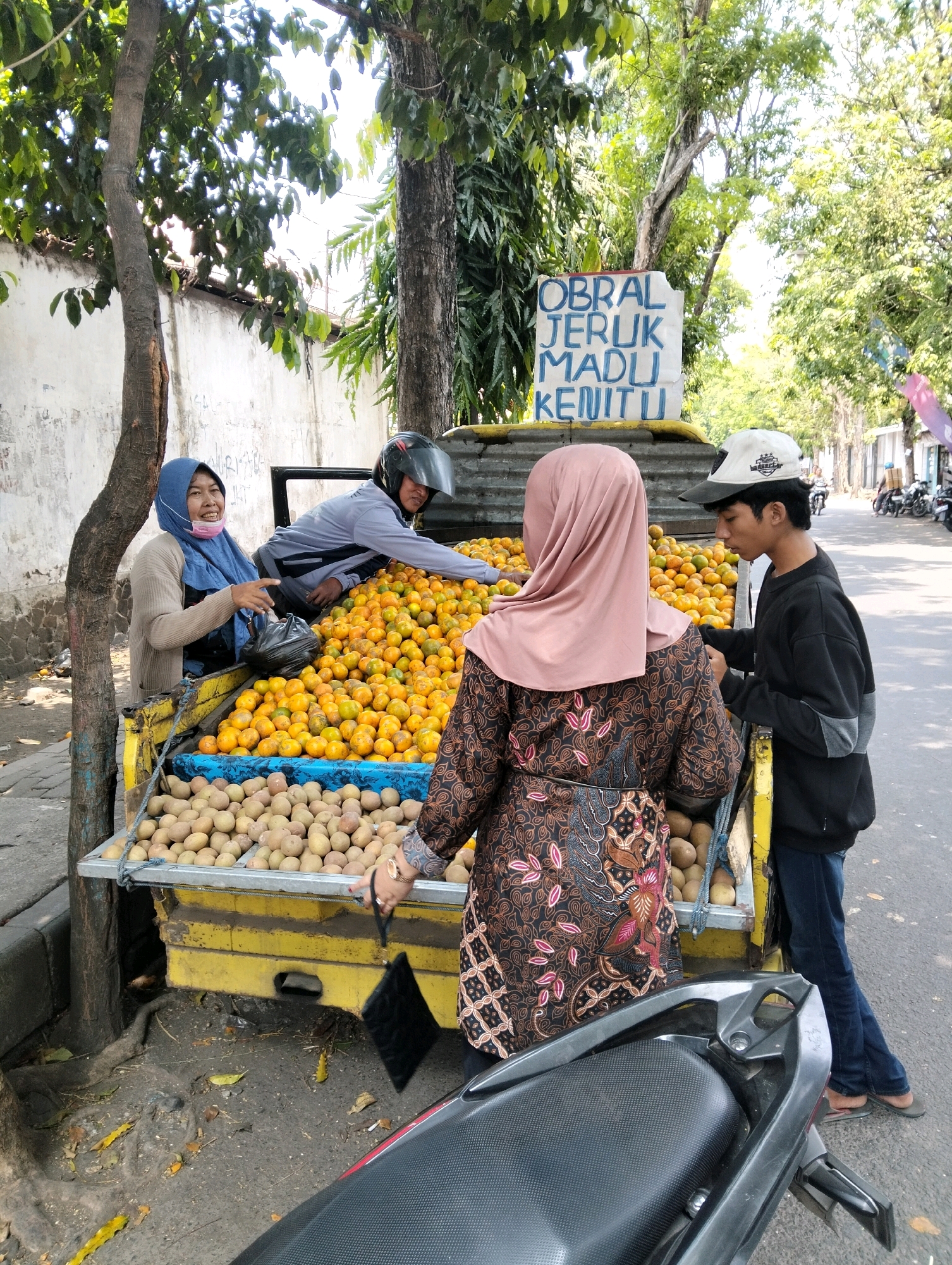 An outdoor medium shot captures a fruit stand on a street, likely in Semampir, Indonesia. A yellow truck bed, serving as the fruit stall, is laden with a large quantity of oranges in the upper portion and smaller, lighter-colored fruits, possibly sapodillas, in the lower portion. Three people are present. A woman wearing a blue hijab and a light-colored cardigan stands to the left, gesturing with her right hand while her left hand holds a black plastic bag. She is smiling and appears to be interacting with a person seated in the truck bed. This seated person, wearing a helmet and a grey long-sleeved shirt, is also looking towards the woman. On the right side of the frame, a young man wearing a white baseball cap and a black long-sleeved shirt is facing the woman and looking down at the fruit. A woman in a patterned brown and pink headscarf and a matching dress is standing in the foreground on the right, facing away from the camera and towards the fruit stand. A dark-colored motorcycle is parked in the lower foreground. In the background, trees line the street, and some buildings are visible. A sign on the truck bed reads "OBRAL JERUK MADU KENITU" in blue lettering.
