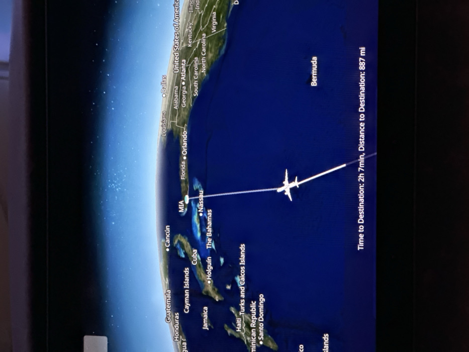 This media file displays an in-flight entertainment screen showing a digital map tracking an airplane's progress. The image is rotated 90 degrees clockwise, so the map appears on its side.
The map illustrates a curved Earth, focusing on the southeastern United States, the Caribbean Sea, and the western Atlantic Ocean. Landmasses are depicted in green and brown, while oceans are dark blue with a lighter blue gradient at the horizon, possibly indicating a nighttime or space view with scattered white dots.
A white airplane icon is visible along a straight white flight path. The origin of the flight appears to be near Florida or the Bahamas, with the current position indicated by the plane icon, and the destination is Bermuda.
Visible text labels on the map include:
- **Countries/Regions:** United States of America, The Bahamas, Guatemala, Honduras, Jamaica, Dominican Republic, Haiti, Turks and Caicos Islands, Cuba.
- **States (USA):** Louisiana, Alabama, Georgia, Florida, Kentucky, Illinois, Indiana, Virginia, North Carolina, South Carolina.
- **Cities/Airports:** Dallas, Atlanta, Orlando, MIA (Miami), Nassau, Cancún, Holguín, Santo Domingo.
- **Islands/Territories:** Bermuda, Cayman Islands.
- **Flight Information:** "Time to Destination: 2h 7min, Distance to Destination: 887 mi".