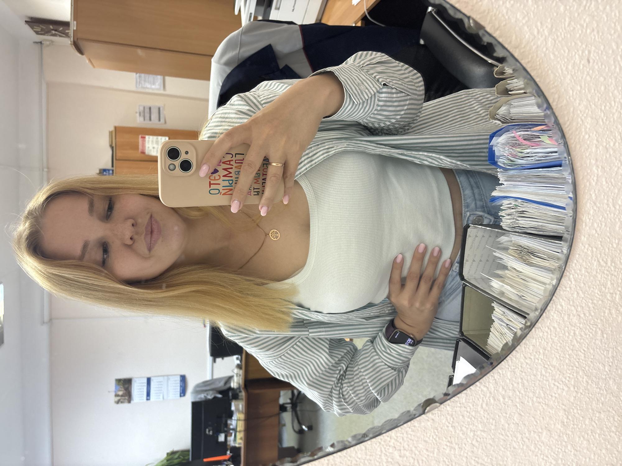 A young blonde woman is taking a mirror selfie in an office environment. She is wearing a light-colored tank top and a vertically striped button-down shirt over it. She has a necklace with a circular pendant and a ring on her right hand. Her phone, with a light-colored case displaying colorful text, is held in her right hand. She is wearing a dark smartwatch on her left wrist and has her left hand on her stomach. The background appears to be an office with desks, chairs, cabinets, and a printer visible in the reflection. The mirror itself has a textured frame and is positioned on a wall with a textured finish. The overall scene suggests a casual moment captured in a workplace. While the city of Заволжье, Russia is mentioned, there are no specific visual cues in the image to confirm this location.