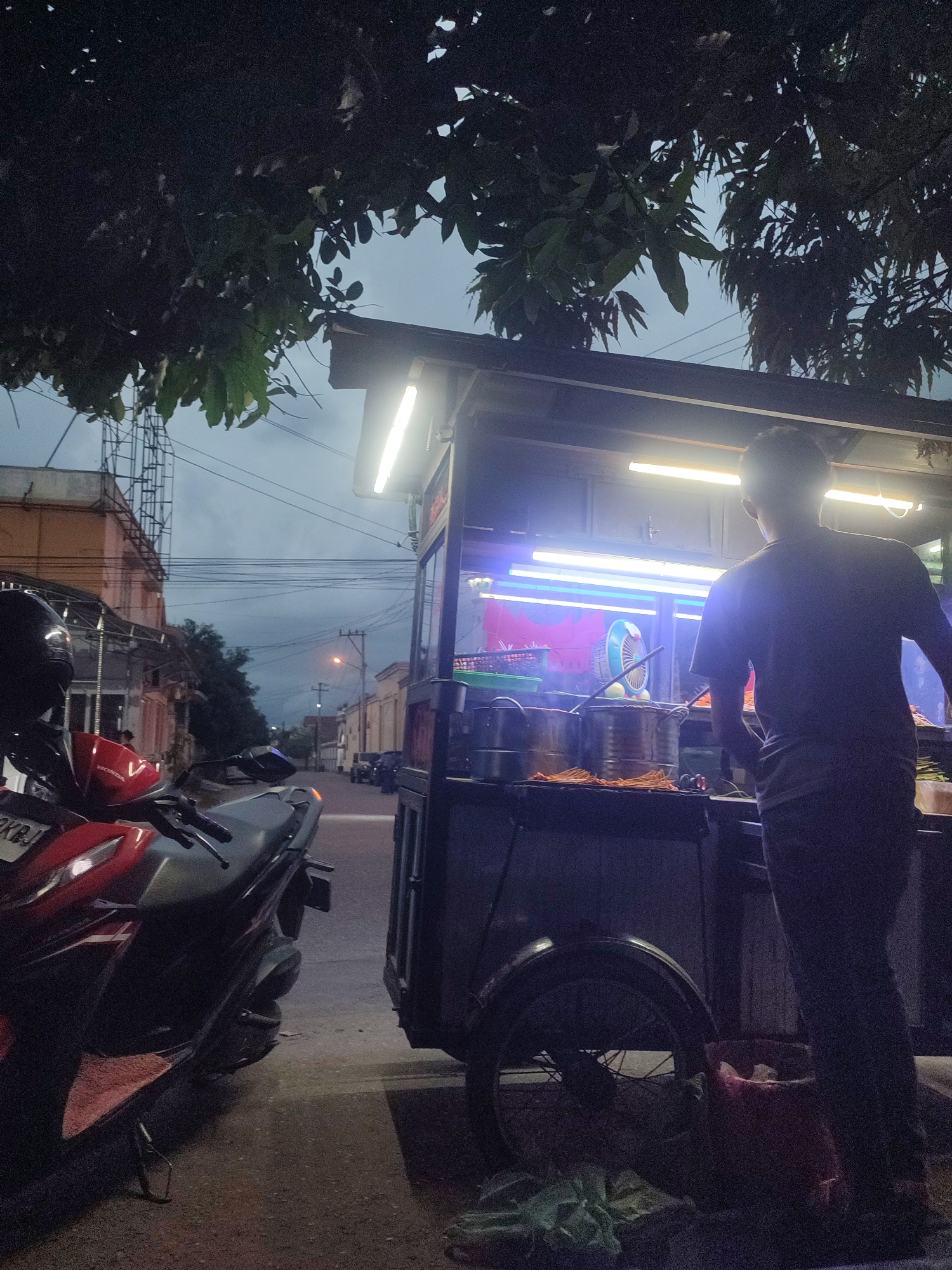 The image shows a street scene in Lhokseumawe, Indonesia at night. A man is standing next to a food cart illuminated by fluorescent lights. The cart appears to be set up for cooking or serving food, as suggested by the presence of pots, utensils, and what seems to be food items. A red and grey motorcycle is parked on the street near the cart. The street is empty except for a few cars parked further down. Buildings line the street, and power lines are visible overhead. Trees with dense foliage are also present. The sky is overcast.