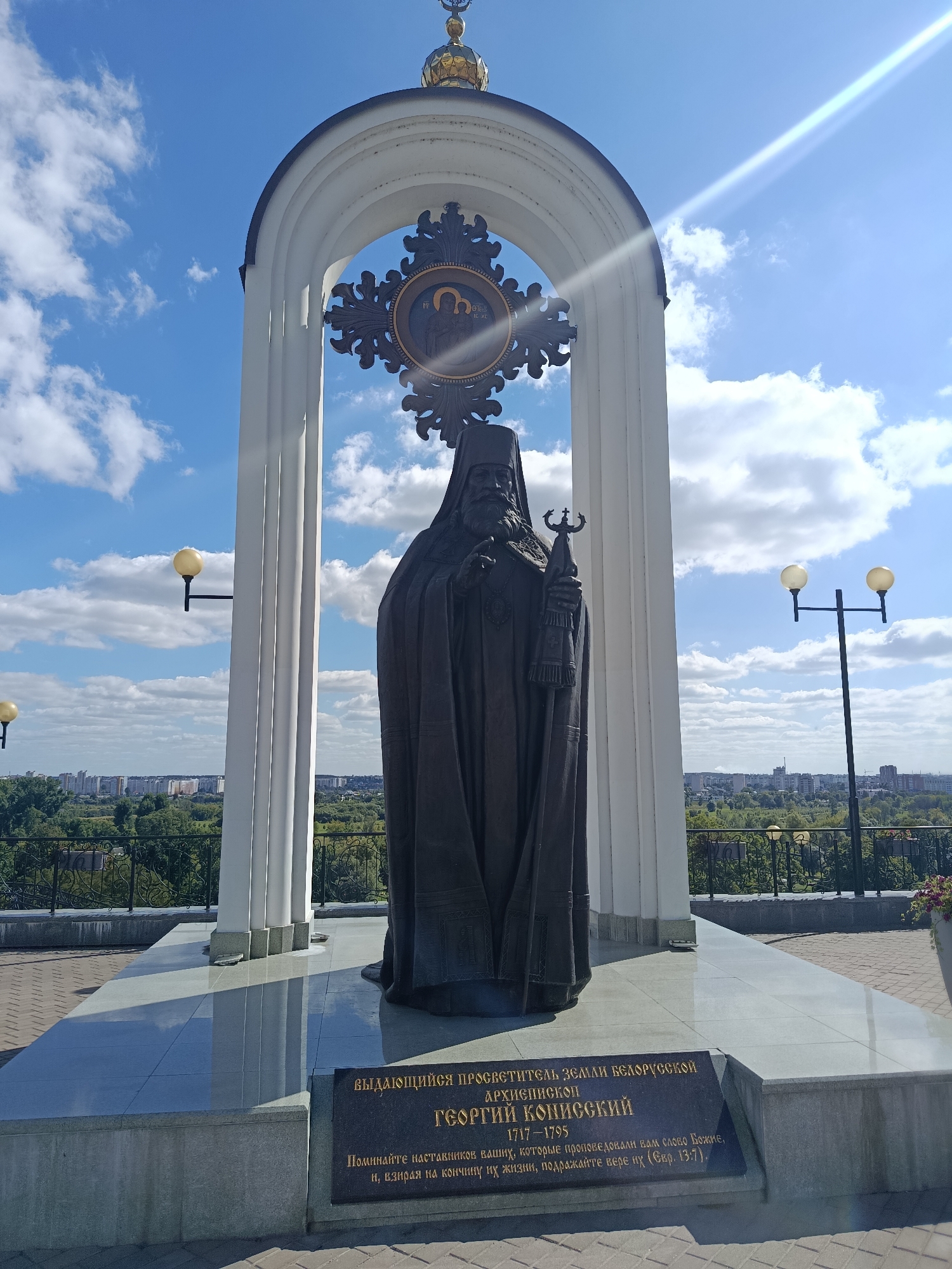 The image shows a statue of a religious figure, identified by an inscription as Archbishop Georgiy Konisskiy, standing in an arched monument in Mahilyow, Belarus. The statue is dark in color and depicts the figure in religious attire, holding a staff. Behind the statue, an ornate cross with religious iconography is visible, framed by the arch. The monument is set against a backdrop of a blue sky with scattered clouds and a panoramic view of the city, featuring buildings, trees, and a distant horizon. The ground in front of the monument is paved. The inscription on the monument provides biographical details and a biblical quote. The scene is illuminated by natural sunlight, casting shadows and creating a bright, daytime atmosphere.