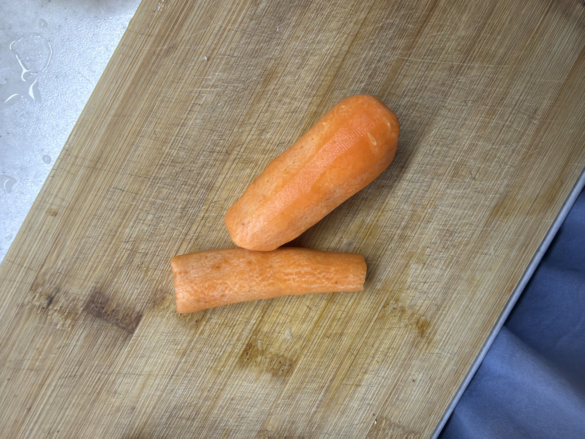 Two peeled orange carrots are positioned on a light-brown wooden cutting board, which exhibits visible knife marks and areas of dark discoloration. One carrot is a relatively complete, peeled specimen, oriented diagonally. The second carrot is a shorter, thicker segment, positioned vertically to the left of the first. In the upper right background, a light-colored, textured countertop surface is partially visible, displaying several distinct water droplets. The lower-left foreground reveals a section of dark blue-grey fabric. The scene is located within a kitchen setting in Chaikovsky, Russia.