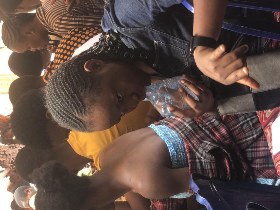 The scene depicts a densely populated indoor or covered gathering at Akpaka Forest Reser, Nigeria. In the foreground, a young female with dark skin, styled cornrow braids, and a denim shirt is observed drinking from a clear plastic water sachet held with both hands. A black smartwatch is visible on her left wrist. To her left, another young female, also with dark skin, is visible from the back, wearing a light blue strapless top with a red and black plaid fabric draped over her waist, and her hair styled in two buns. Both individuals are seated on blue plastic chairs. The background reveals a crowd of numerous individuals, predominantly young adults, seated in rows. Clothing patterns include solid yellow, polka dots, and stripes. Some individuals in the background appear to be interacting with mobile phones. The overall environment suggests an organized assembly or event.