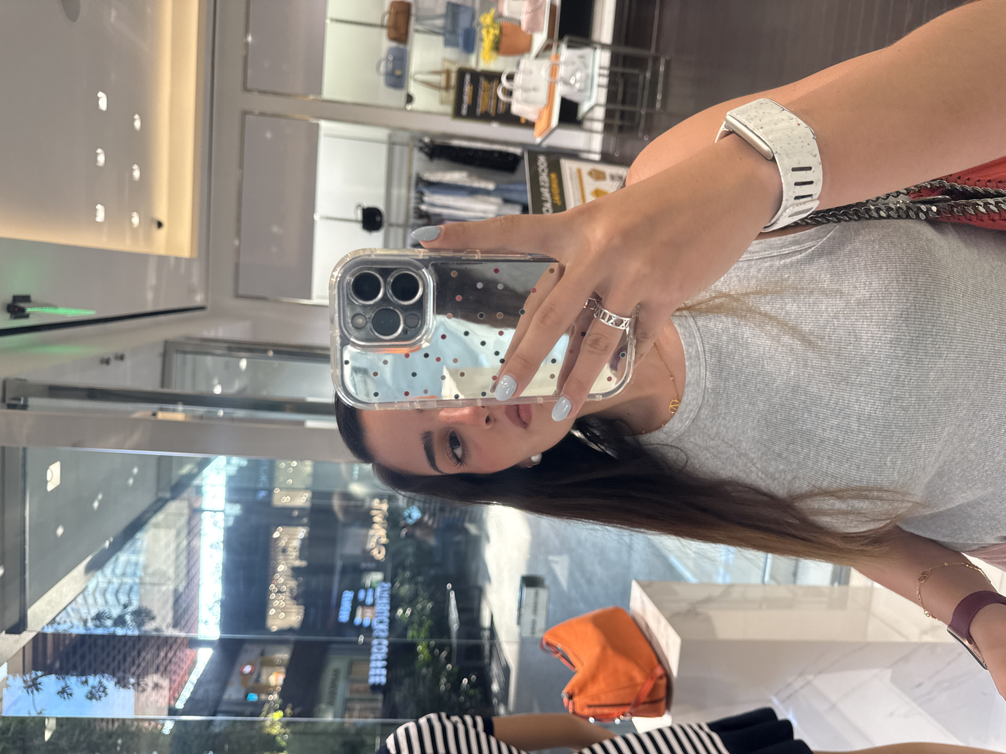 A woman takes a selfie inside a store, likely in Puebla, Mexico. She is holding a smartphone with a clear case decorated with polka dots. The phone's camera is visible. The woman has long brown hair, light blue nail polish, and wears a silver ring and a gold necklace. She is wearing a light gray shirt. A white smartwatch is on her left wrist. She is standing in front of a glass window that reflects an outdoor scene with buildings and trees. Inside the store, there are shelves with handbags and clothing. An orange handbag is on display. The interior of the store appears well-lit with modern fixtures.