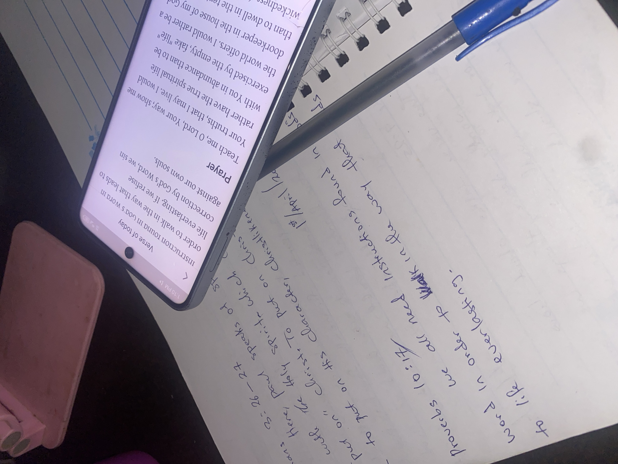 A top-down, close-up shot shows a person's handwritten notes alongside a mobile phone displaying a devotional prayer. The notes, written in blue ink on white paper, include a Bible verse reference (Proverbs 10:17) and the date "1st / April /20". The prayer on the phone discusses walking in God's word and seeking correction. A blue pen rests on top of the paper and the phone, along with a pink phone stand. The scene is set indoors with a dark surface visible in the background.