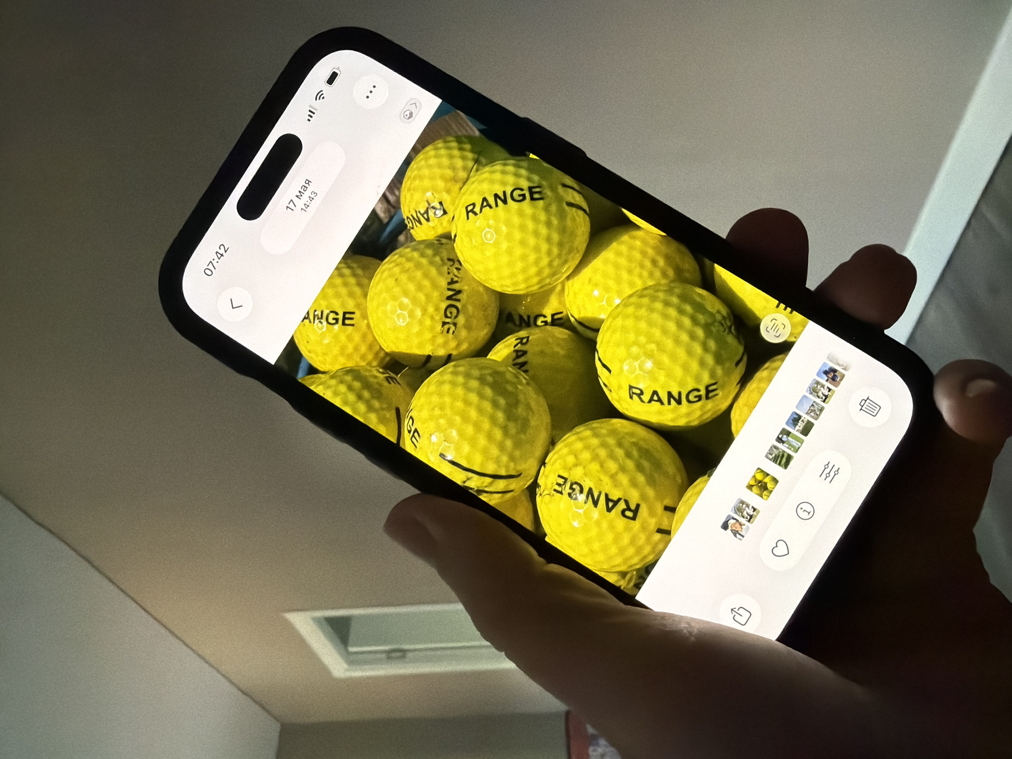 A hand is holding a smartphone displaying a picture of yellow golf balls. The golf balls are the primary subject of the image on the phone screen and appear to be from a driving range. The phone is held at an angle, obscuring part of the background. The background of the original image shows an indoor setting with a wall and ceiling. There is no visible information within the image to suggest a location in Malinovka, Russia.