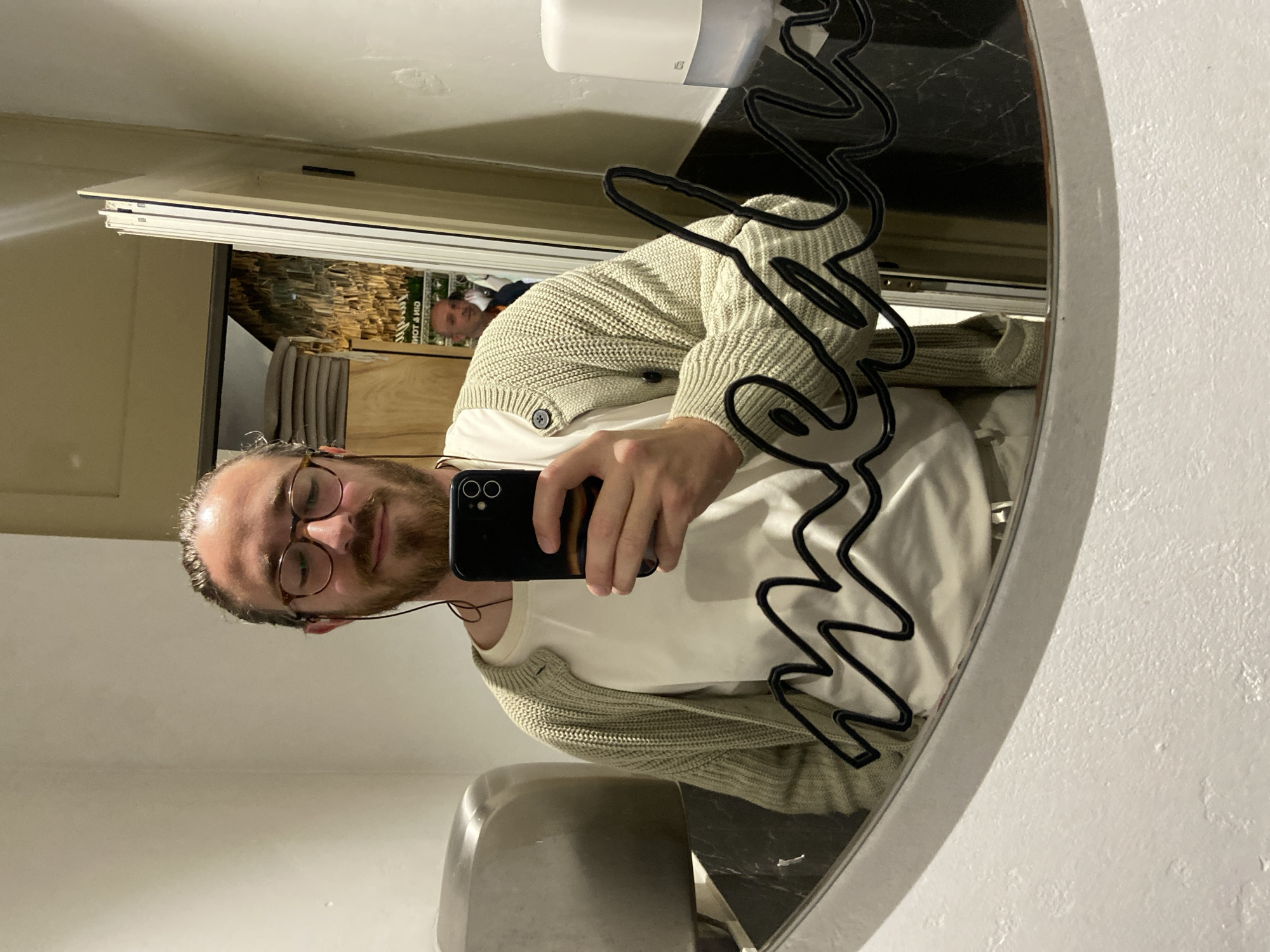 A Caucasian male with a beard and wearing glasses is taking a selfie in a mirror. He is wearing a white T-shirt and a light green cardigan. In the reflection in the mirror, behind him, there is another man visible. The background appears to be a bathroom. The specific location of Petrovac centar, Montenegro, cannot be confirmed from the visual data.