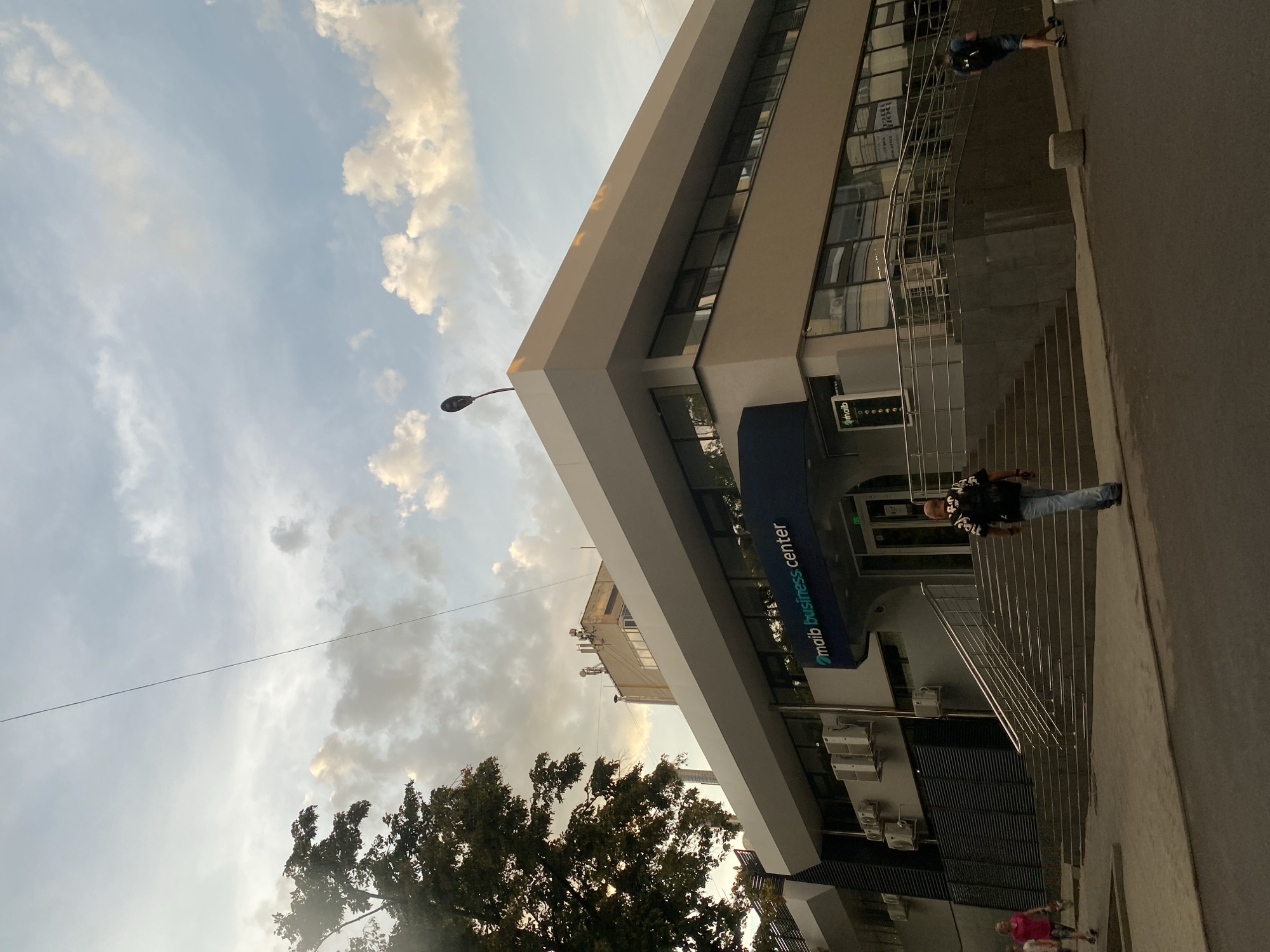 The image shows the MAIB Business Center, a modern building located in Chișinău, Moldova. The building features a sleek, contemporary design with large windows and a prominent blue sign displaying the business center's name. In front of the building, a wide set of stairs leads to the entrance, flanked by metal railings. Several individuals are visible, some ascending or descending the stairs, while others walk along the sidewalk. The sky above is filled with dramatic clouds, suggesting either a late afternoon or early morning setting. The overall atmosphere is one of urban activity, with the business center serving as a focal point within the cityscape.