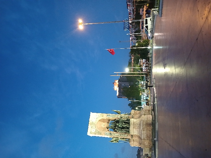 The image shows the Republic Monument in Taksim Square, Istanbul, Türkiye (latitude: 41.037, longitude: 28.985). The monument is a large, rectangular structure made of stone, with a series of statues of people on top of it. The monument is lit up, and there are a few people walking around it. In the background, there are some trees, buildings, a Turkish flag, and a street lamp. The sky is a dark blue, and there are a few clouds in the sky. The square is empty and paved. A white van is parked at the right edge of the square. A tall, thin building with lights on can be seen in the background. The scene is lit by street lights and the lights on the monument. There are a few people standing in front of the monument.