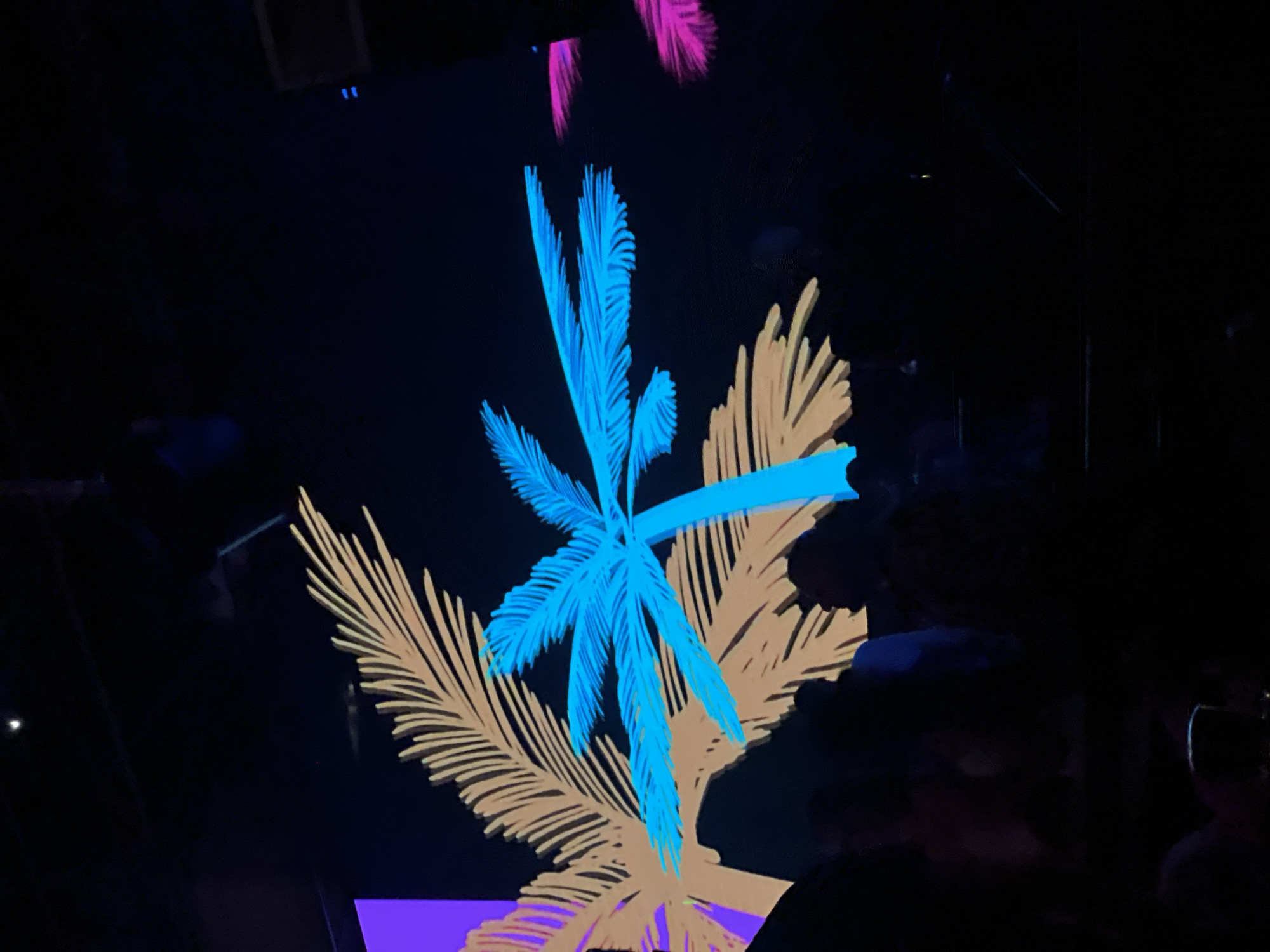 This image depicts a dark indoor environment, likely a club or concert venue, with a large projection screen displaying stylized palm tree graphics. Two prominent palm tree illustrations are visible: one in a tan or light brown color with a purple section on the left side, and another in a vibrant cyan or light blue color. The cyan palm tree is larger and positioned in the foreground, overlapping the tan one. Smaller, fragmented palm frond graphics in hot pink are visible on the right side of the screen.
Below the projection screen, the silhouettes of several people are visible, suggesting an audience. The lighting is dim, with the colorful graphics on the screen being the primary light source. The overall atmosphere appears to be one of entertainment or a nightlife event. The location is stated to be Sharm El Sheikh, Egypt, which is a popular tourist destination known for its resorts, beaches, and nightlife.