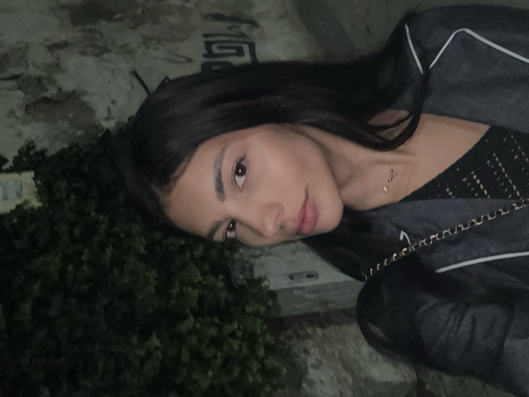 A young woman with dark hair and light skin is the focal point of the image. She is wearing a dark top and a jacket with a white trim. A delicate necklace is visible around her neck. The woman is positioned at an angle, looking towards the camera. The background consists of a stone wall with some graffiti and dense foliage, suggesting an outdoor setting. The image was taken in an urban area during nighttime. Although it is not clearly identifiable, the location is presumed to be in the city of Plovdiv, Bulgaria.