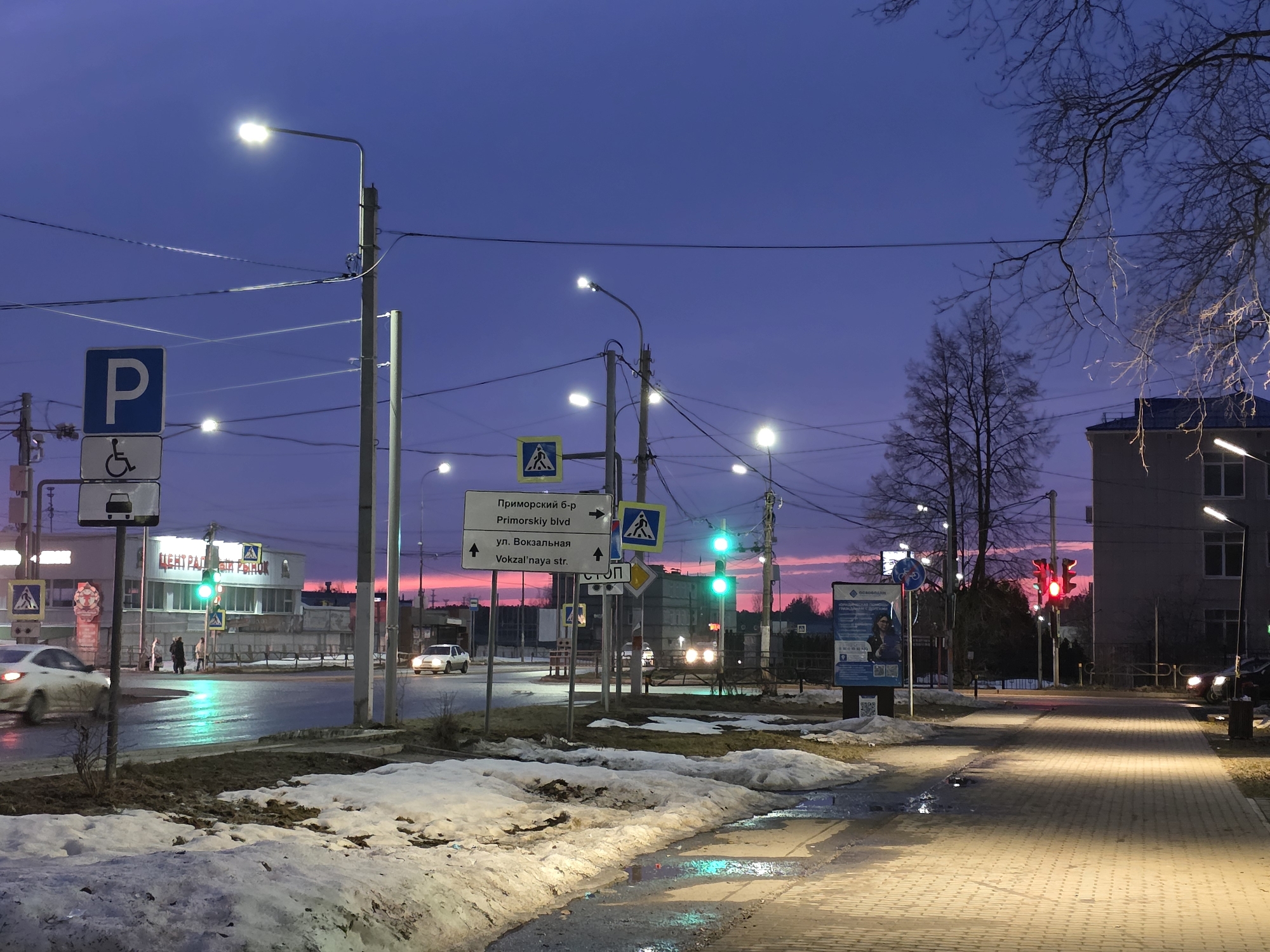 The image shows a street scene in what appears to be the city of Chaikovsky, Russia, at dusk or dawn. The sky is a mix of blue and pink hues. The photograph is taken from a sidewalk, which is paved with rectangular stones. Patches of melting snow are visible on the side of the road.
Several street signs and traffic lights are present. One sign indicates "Primorskiy blvd" and "Vokzal'naya str", likely street names. There's also a parking sign, and several crosswalk signs with pedestrian crossings. The traffic lights show both green and red signals.
Buildings can be seen in the background, including one identified as "ЦЕНТРАЛЬНЫЙ РЫНОК" (Central Market). Cars are visible on the road, some with headlights on. Bare tree branches extend into the sky. Streetlights illuminate the scene.