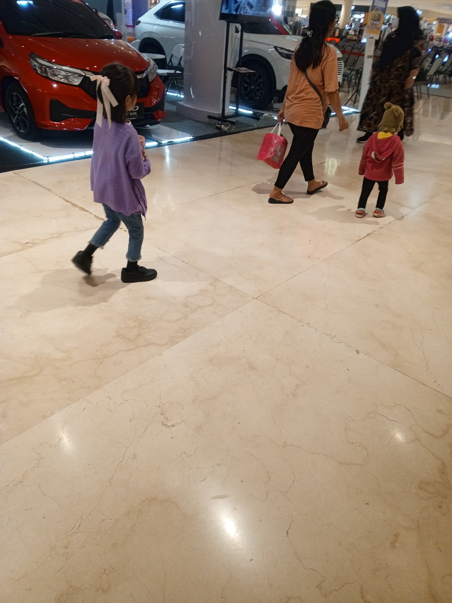 The image depicts a bright, indoor environment, likely a shopping mall, with a polished marble floor. In the foreground, a young girl wearing a purple sweater and blue jeans walks away from the camera, her dark hair tied with a light-colored bow. To her right, a woman in an orange shirt and black pants walks towards the camera, carrying a pink shopping bag. Behind them, another woman in a patterned dress walks away from the camera. A small child in a red jacket and green hat is walking ahead of the woman in the orange shirt. In the background, a red car and a white car are displayed. The scene is well-lit, suggesting artificial lighting typical of indoor retail spaces. Based on the context of the cars being displayed, this scene likely takes place at a car dealership showroom within a shopping mall in Cibinong, Indonesia, possibly at Cibinong City Mall.