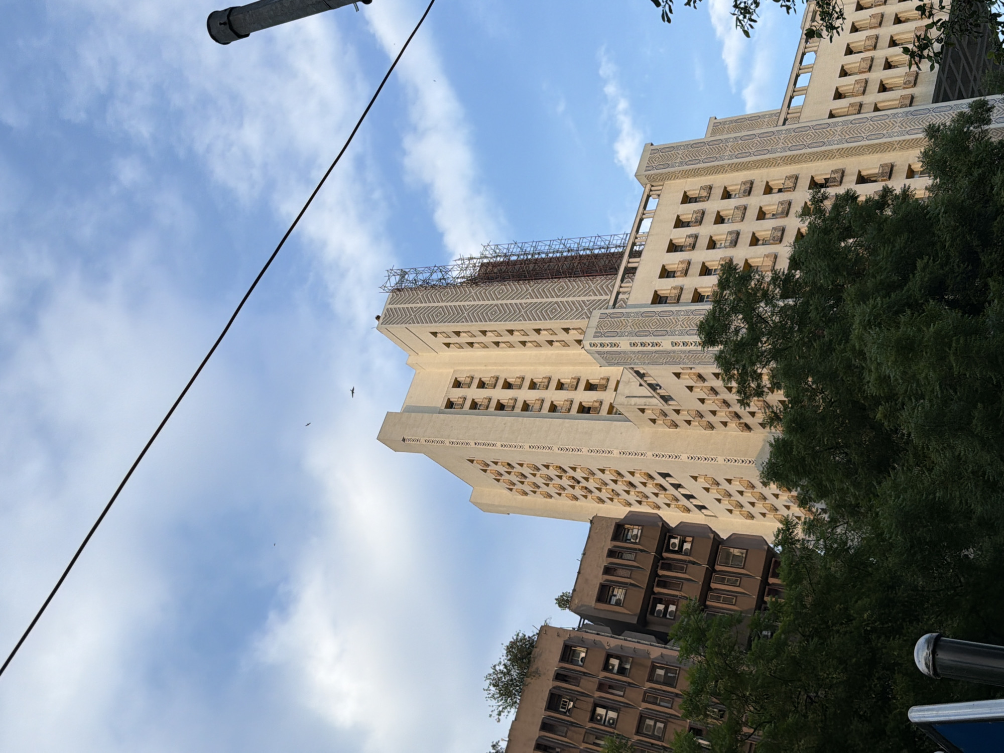 A low-angle outdoor shot captures the upper floors of several buildings against a cloudy blue sky. The central and tallest building is a pale, cream-colored structure with numerous small balconies and decorative geometric patterns along its facade. Scaffolding is erected on the upper section of this building, suggesting ongoing construction or renovation. To the left, two older, darker brown buildings are visible, also with multiple windows and balconies, some of which appear to be fitted with air conditioning units. Lush green trees obscure the lower portions of the buildings and extend upwards, partially framing the structures. A thick black cable stretches diagonally across the upper part of the frame, and a dark, cylindrical object, possibly a chimney or a part of a utility pole, is visible on the far right. The image appears to be taken in a densely built urban environment. The architectural style and the presence of trees suggest this could be an older, more established area within New Delhi, India.