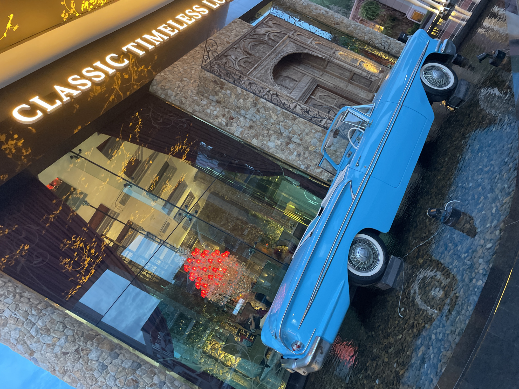 The image shows a vibrant blue classic convertible car parked in a shallow water feature in front of a building with a stone facade and large glass windows. A crystal chandelier with red lights is visible through the window, along with reflections of the sky and building interior. Above the entrance, illuminated white letters spell out "CLASSIC TIMELESS LO". The car's wheels are resting on dark rectangular blocks submerged in the water, which is filled with small pebbles. The scene appears to be an outdoor display, possibly a hotel or resort in Pattaya City, Thailand.