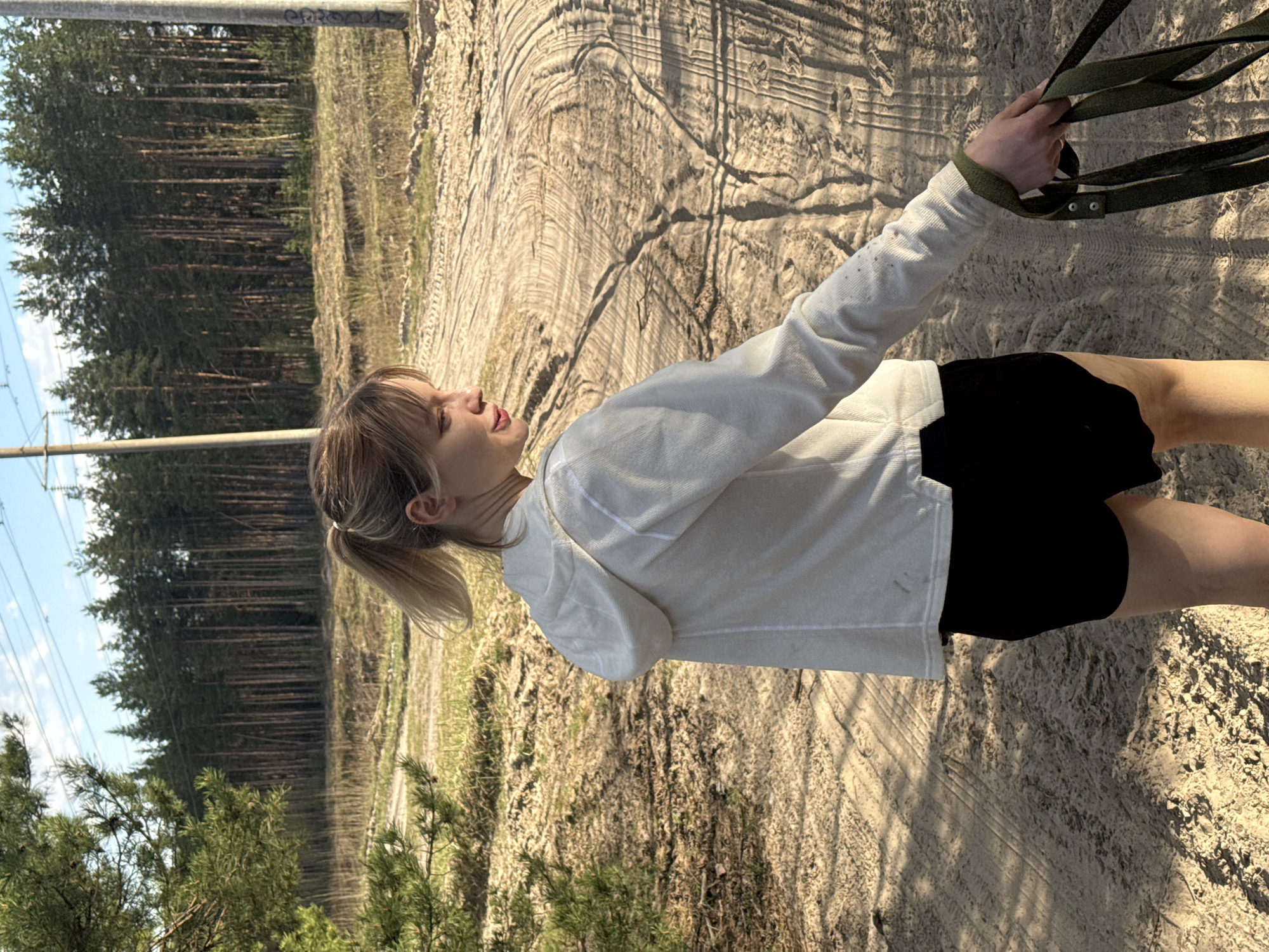 A woman with blond hair pulled into a ponytail is standing on a dirt path with tire tracks and holding green straps in her right hand. She is wearing a white hoodie and black shorts. The background includes a forest of pine trees, a power line, and a sandy area. The exact location in Nizhny Novgorod, Russia, cannot be determined from this image.