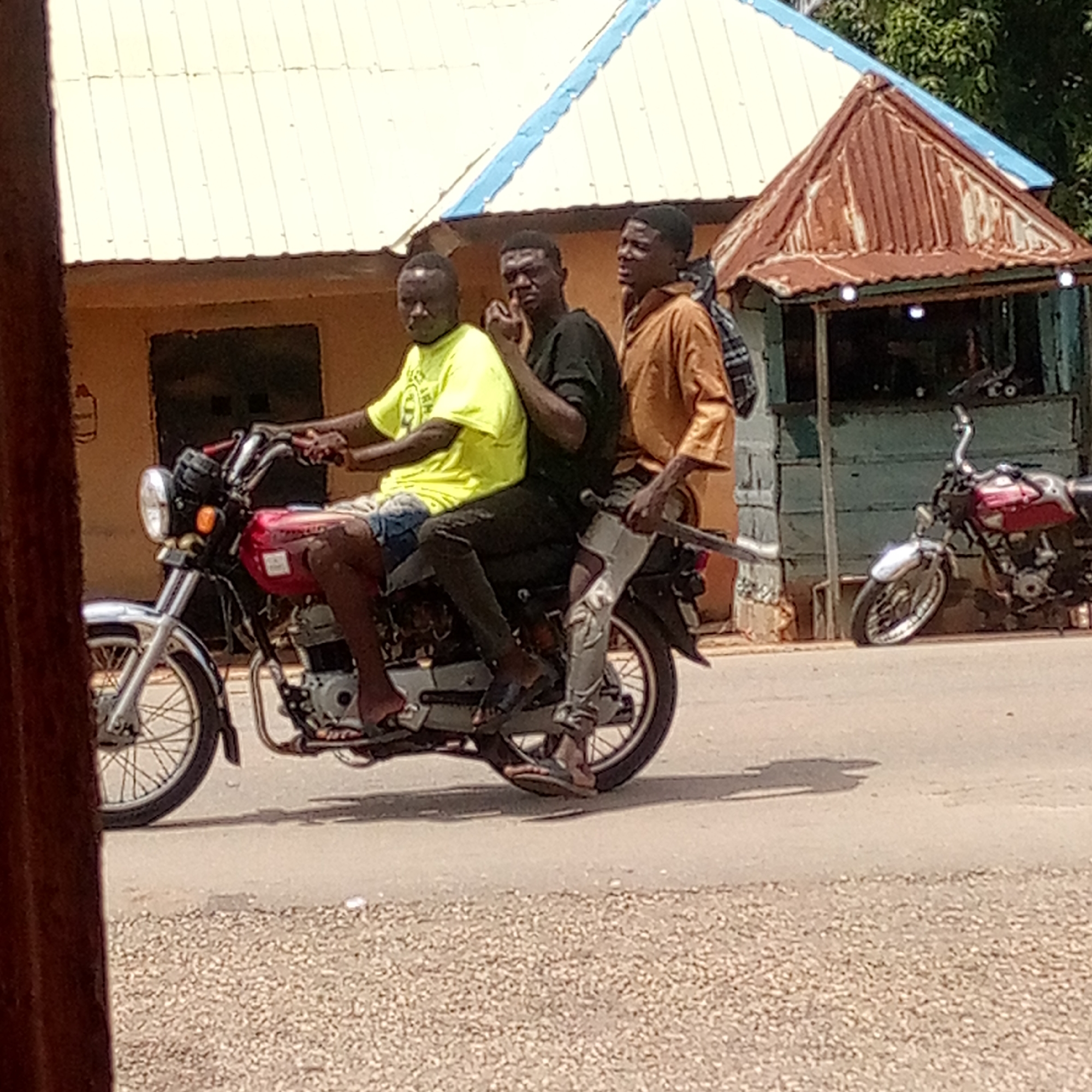 The image captures three young men riding a red and silver motorcycle on a paved road in Mararaba, Nigeria, under bright daylight.
The driver, positioned at the front, wears a bright yellow t-shirt and blue shorts, with flip-flops on his feet. The middle passenger is dressed in a dark t-shirt and dark pants, also wearing flip-flops, and is seen with his hand near his face. The rear passenger, wearing a tan long-sleeved shirt and dark pants, carries a backpack and also wears flip-flops. The motorcycle features a red fuel tank, silver handlebars, and spoked wheels, appearing to be a common model for local transport.
The scene suggests a semi-urban or rural environment. In the background, to the left, is a building with a light-colored corrugated metal roof, possibly white or cream, and blue trim. To the right, a smaller structure, resembling a kiosk or stall, has a distinct rusty brown corrugated metal roof. Green foliage is visible behind this smaller building. Another red motorcycle is parked near the right-side structure. The strong sunlight indicates a clear, sunny day, with visible shadows cast on the road. No legible text is visible on any subjects or objects. The image depicts a common mode of transportation and everyday activity in the region.