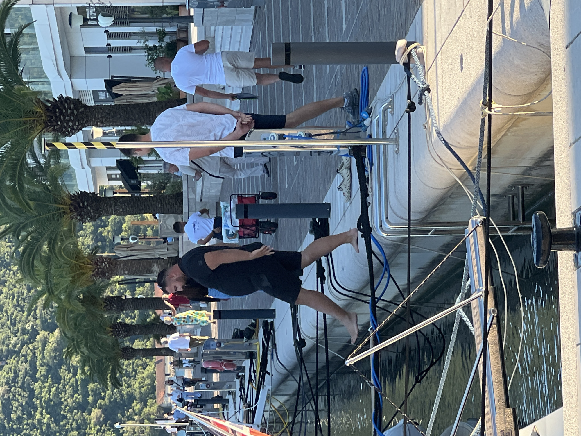 The image depicts a sunny day in Kumbor, Montenegro, with several people visible on a waterfront promenade and near a docked boat. The setting features modern architecture, palm trees, and the water. One man in the foreground is wearing a black t-shirt and shorts and is standing with his left leg extended, appearing to be in motion or posing. Behind him, another man in a white t-shirt and light shorts is walking. Further back, a group of people are gathered, some of whom are dressed in white. The water is calm, reflecting the light. The scene suggests a leisurely atmosphere, possibly a tourist area. There is no visible text in the image.