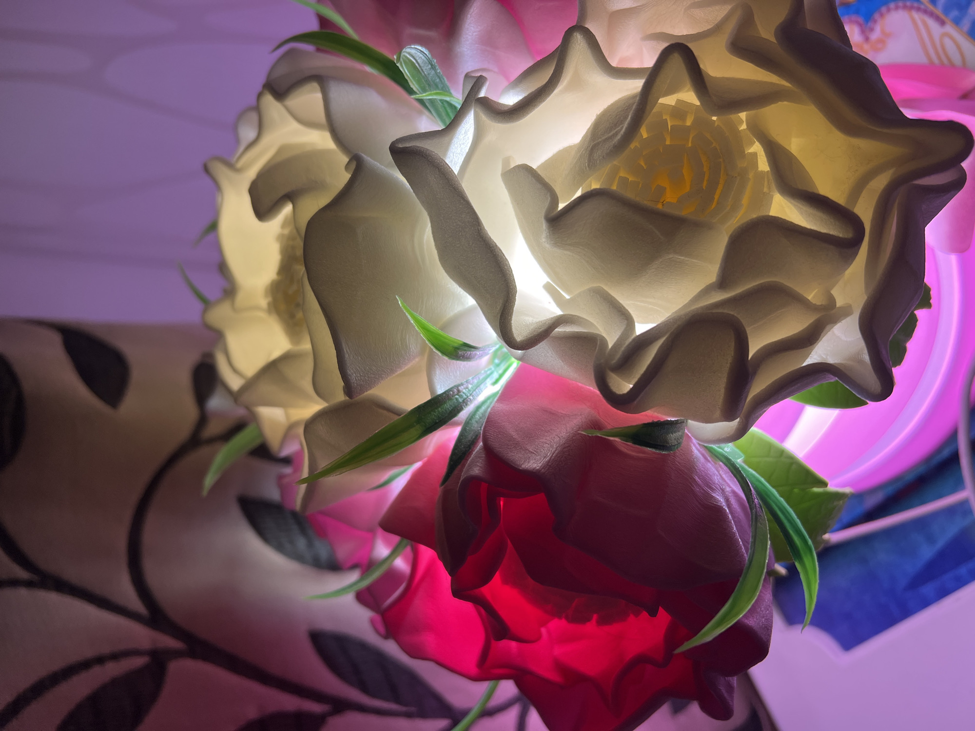 The image captures a close-up of an illuminated decorative floral arrangement, likely an indoor light fixture, set in Perm, Russia. The primary subjects are several artificial flowers, glowing softly from within.
Dominating the center and upper portions of the frame are two large, white or cream-colored flowers. Their translucent, intricately sculpted petals reveal a textured, almost cellular core that emits a bright, warm light. Below these, one or more red and pink flowers are visible, also glowing warmly from within, casting a deep crimson and magenta light. Slender green leaf-like accents are interspersed among the petals of the flowers.
The background on the left features a softly blurred brown fabric adorned with a dark, swirling floral or abstract pattern, suggesting upholstery or a curtain. To the right, abstract shapes in shades of purple, pink, and blue hint at other components of the light fixture or surrounding decor. The overall lighting is soft and ambient, creating a serene and cozy atmosphere, suggesting the image was taken indoors, possibly in the evening. No specific activity or event is depicted, and no text is visible.