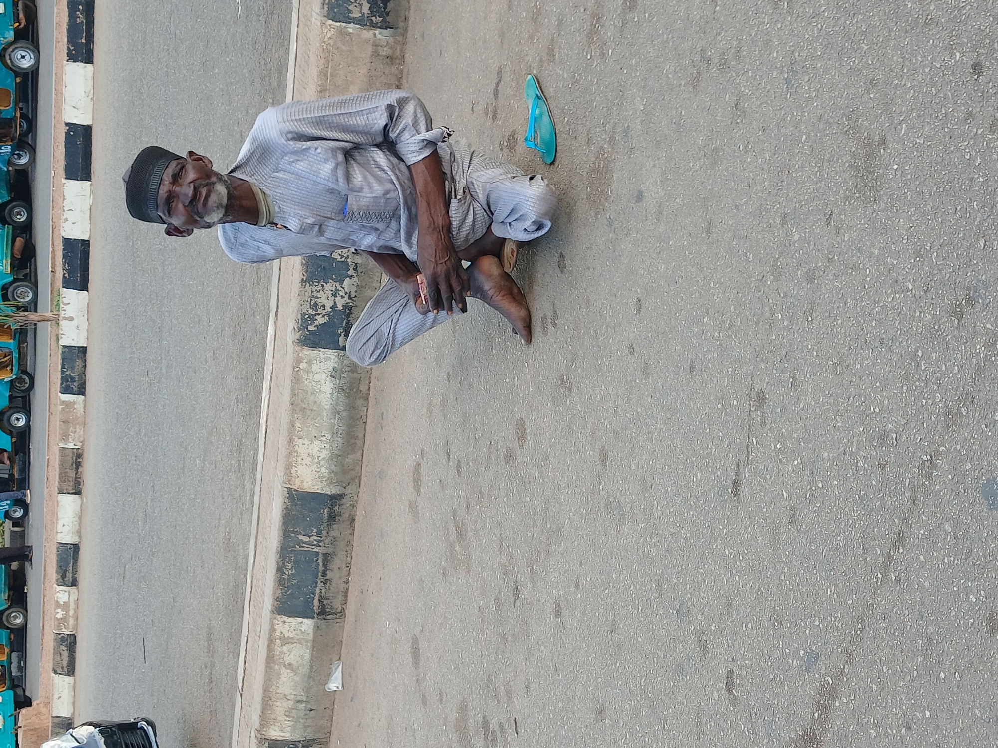 An elderly man, wearing a grey traditional outfit and a black cap, sits on the asphalt next to a curb in Barakin Lerawi, Nigeria. He is looking towards the camera with a slight smile. A single blue flip-flop is visible on the ground to his right. The background shows a paved road with a few parked tricycles.
