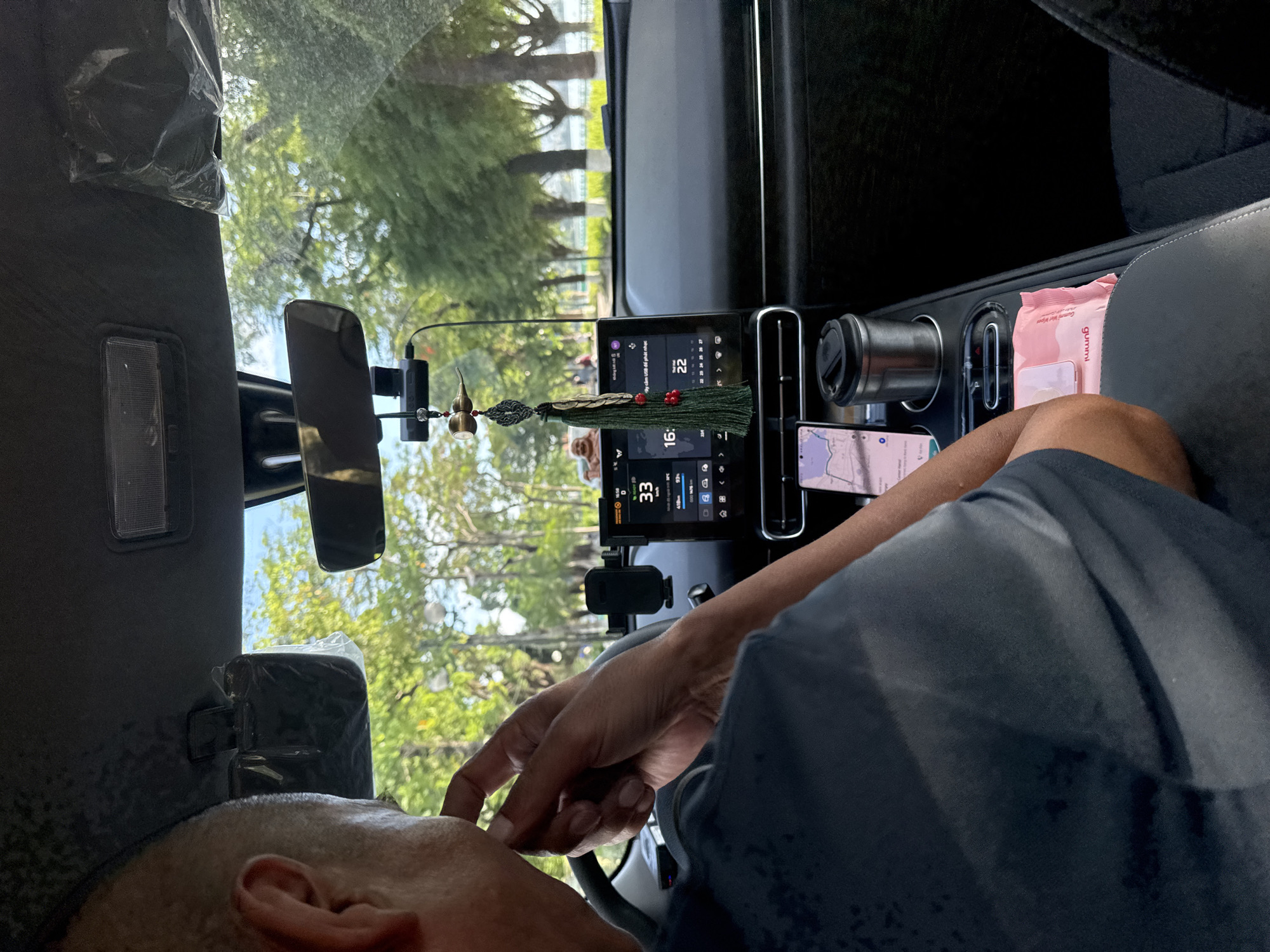 The image shows the interior of a car, likely in Hanoi, Vietnam, as indicated by the text on a wet wipe packet. The scene is viewed from the perspective of a passenger in the back seat, looking towards the dashboard and the driver.
The primary subjects are the driver, whose head and shoulder are visible in the foreground, and the car's dashboard area. The driver is wearing a dark t-shirt and appears to be interacting with something on the dashboard, possibly the screen or a device mounted there. The dashboard features a large touchscreen display showing a navigation map, weather information, and vehicle speed (33 km/h). There are also a car vent, a cup holder with a cup, a phone mounted on a stand displaying a map, and a pack of "gummi" wet wipes. A decorative tassel hangs from the rearview mirror.
The setting appears to be daytime, with natural light filtering in through the windshield, illuminating the trees and greenery visible outside. The weather seems to be clear and warm, judging by the foliage and the presence of the driver's casual attire. The driver's focus seems to be on the dashboard display, and there are no strong emotional cues.
Visible text includes "gummi" on the wet wipe packet and some Vietnamese text on the car's infotainment screen, which appears to relate to navigation and vehicle information. The overall impression is a candid snapshot of a moment inside a car during a drive.