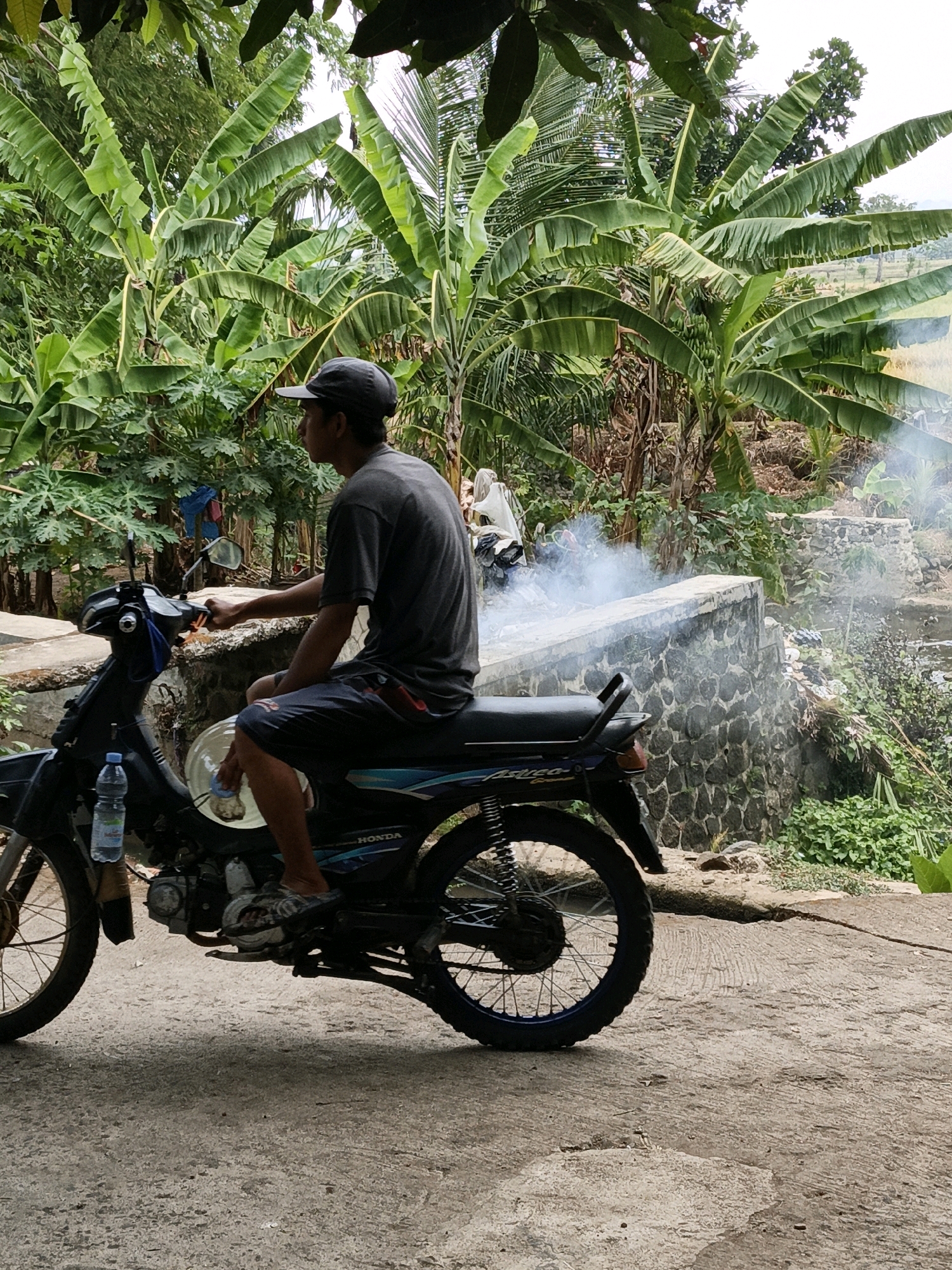 In Mangli, Indonesia, a man wearing a dark grey t-shirt, dark shorts, and a grey baseball cap sits astride a black motorcycle. The motorcycle has a blue stripe along its side panel. A partially consumed bottle of water is attached to the handlebars. The man appears to be cleaning a circular white object positioned on the motorcycle's frame. Smoke billows from behind a stone wall in the background, suggesting a fire or exhaust. The environment is lush with abundant banana trees and other vegetation, and the ground is a rough, paved surface.