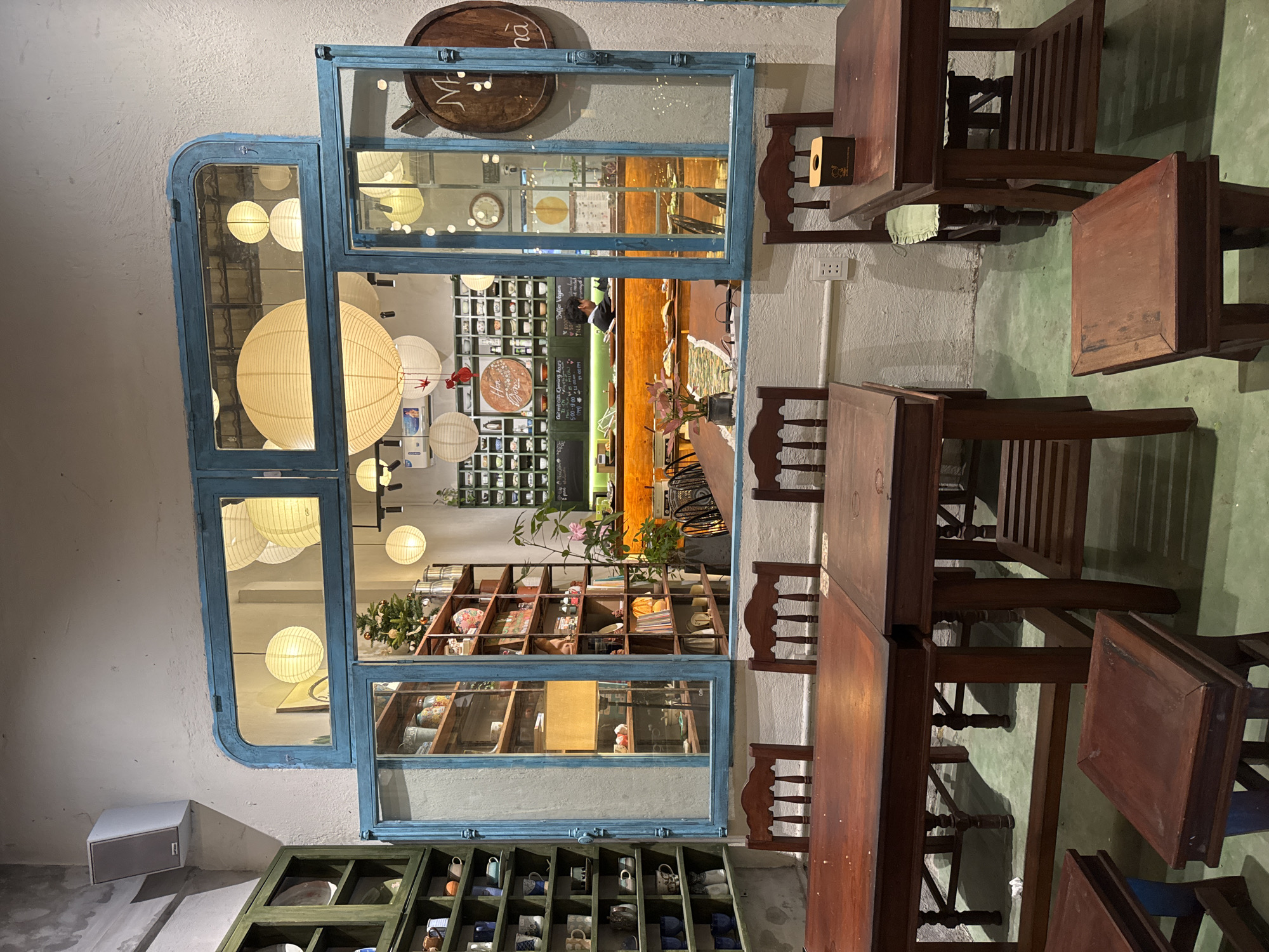 The image shows an interior shot of a cafe or restaurant in Đà Nẵng, Vietnam. The foreground features several wooden tables and chairs arranged in a seating area. A vintage-style, blue-framed window or partition is positioned against the wall, providing a view into another part of the establishment. Through the window, one can see a display of various items on shelves, including books, pottery, and decorative objects. Paper lanterns are visible hanging from the ceiling in the background. There is a counter with a server behind it. The environment suggests a cozy, bohemian atmosphere. The lighting is warm.