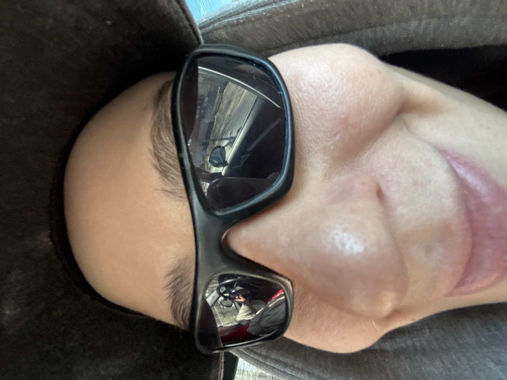 A close-up, high-angle selfie shows a person's face, rotated 90 degrees clockwise, likely taken inside a vehicle during daytime. The person is wearing dark, rectangular-framed sunglasses that cover their eyes. Their forehead, eyebrow, nose, and part of their mouth and cheek are visible. A dark, possibly hooded, garment covers the top and left side of the frame, bordering the person's head.
The lenses of the sunglasses offer reflections of the immediate surroundings. In the upper left lens (right side of the image as rotated), a clear reflection shows the interior of a vehicle, including what appears to be a dashboard or interior panel, and potentially a part of a window or windshield. The lower right lens (left side of the image as rotated) also reflects the vehicle's interior, showing a more detailed view with what might be part of a steering wheel or other vehicle controls, and possibly a glimpse of another person or objects further inside the car.
The person's skin appears fair, with some minor pores visible on the nose and cheek. Their eyebrow is dark and defined. The mouth is slightly open, revealing the upper lip. No specific identifying features of Krasnoyarsk, Russia, are discernible from the reflections or the person's immediate surroundings. There is no visible text in the image.