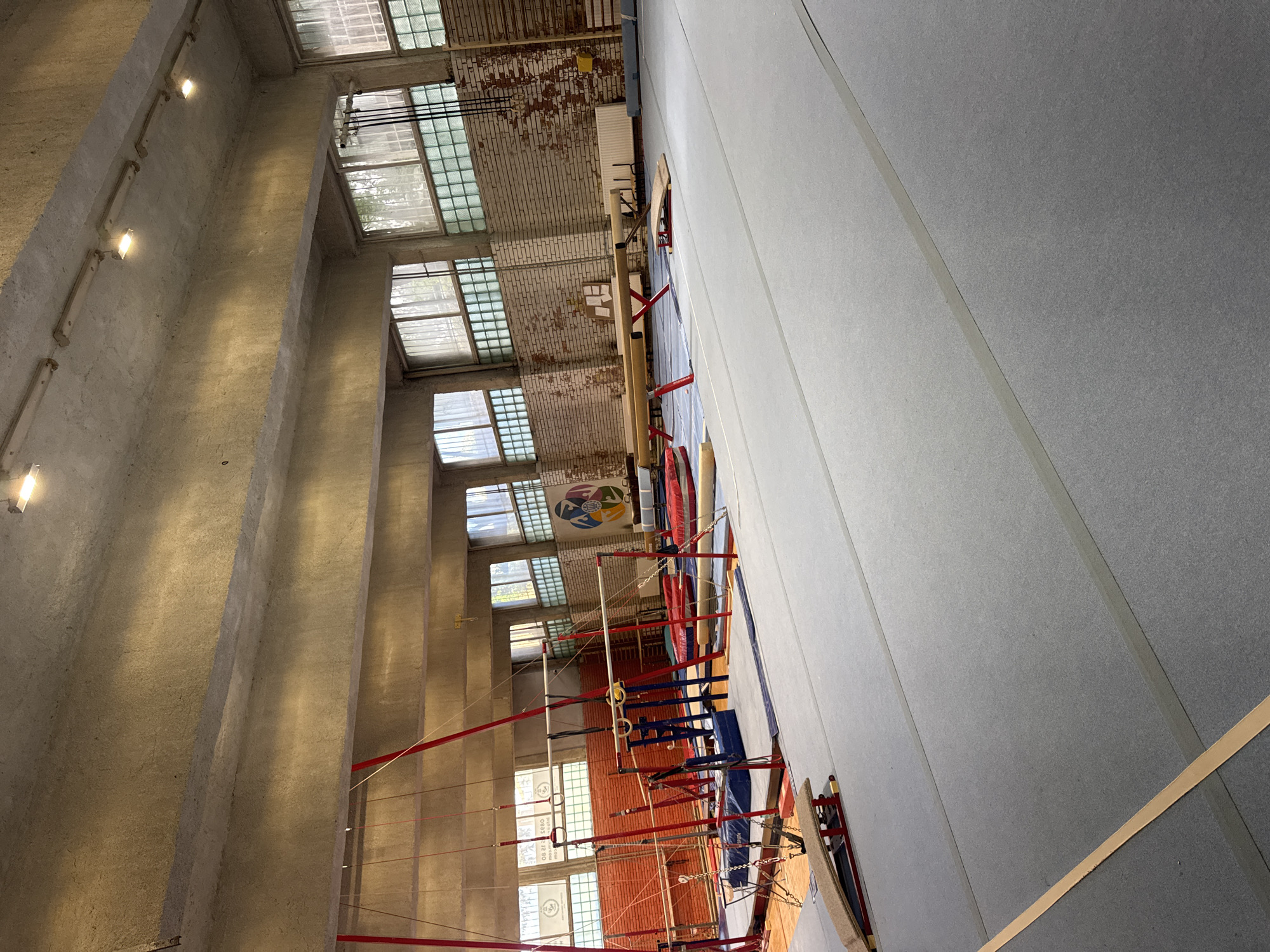 A gymnastic hall, possibly in Sofia, Bulgaria, based on the symbol on the wall, is depicted. The scene is dominated by tall, reinforced concrete pillars with rows of lights attached to them, parallel to an exterior wall with multiple windows. Gymnastic equipment, including rings, bars, and possibly ropes, is placed on a grey floor, with some equipment set up with red and blue supports. The walls are partially brick, with some sections painted light grey. The hall appears to be spacious and well-lit.